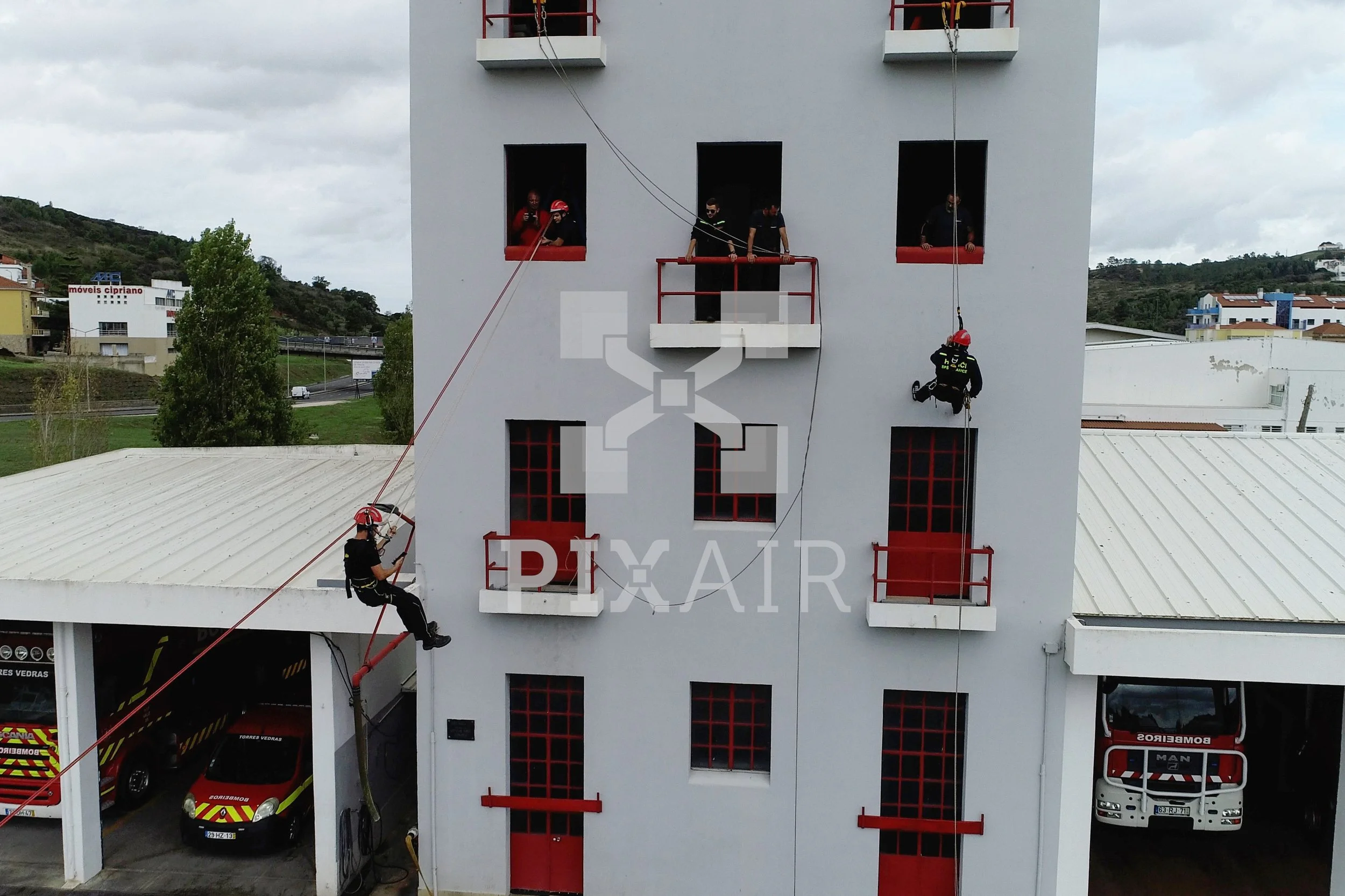 Bombeiros realizando treinamento de resgate e salvamento em um prédio de vários andares, com várias pessoas observando de janelas e uma pessoa sendo içada por corda de resgate.