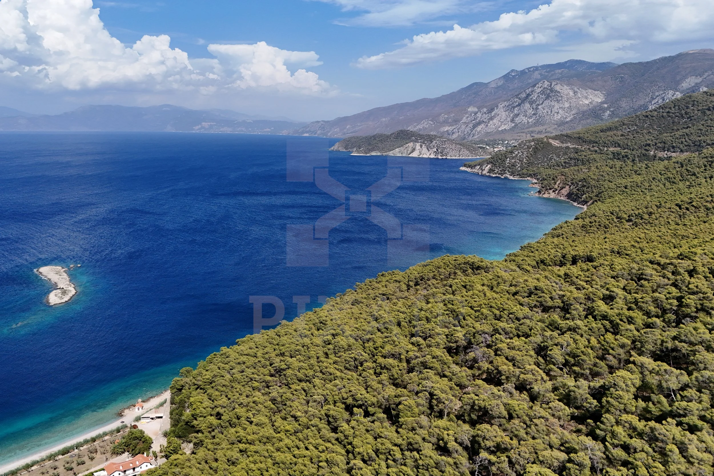 Cenário de paisagem com mar azul, floresta densa na margem e montanhas ao fundo sob céu parcialmente nublado.