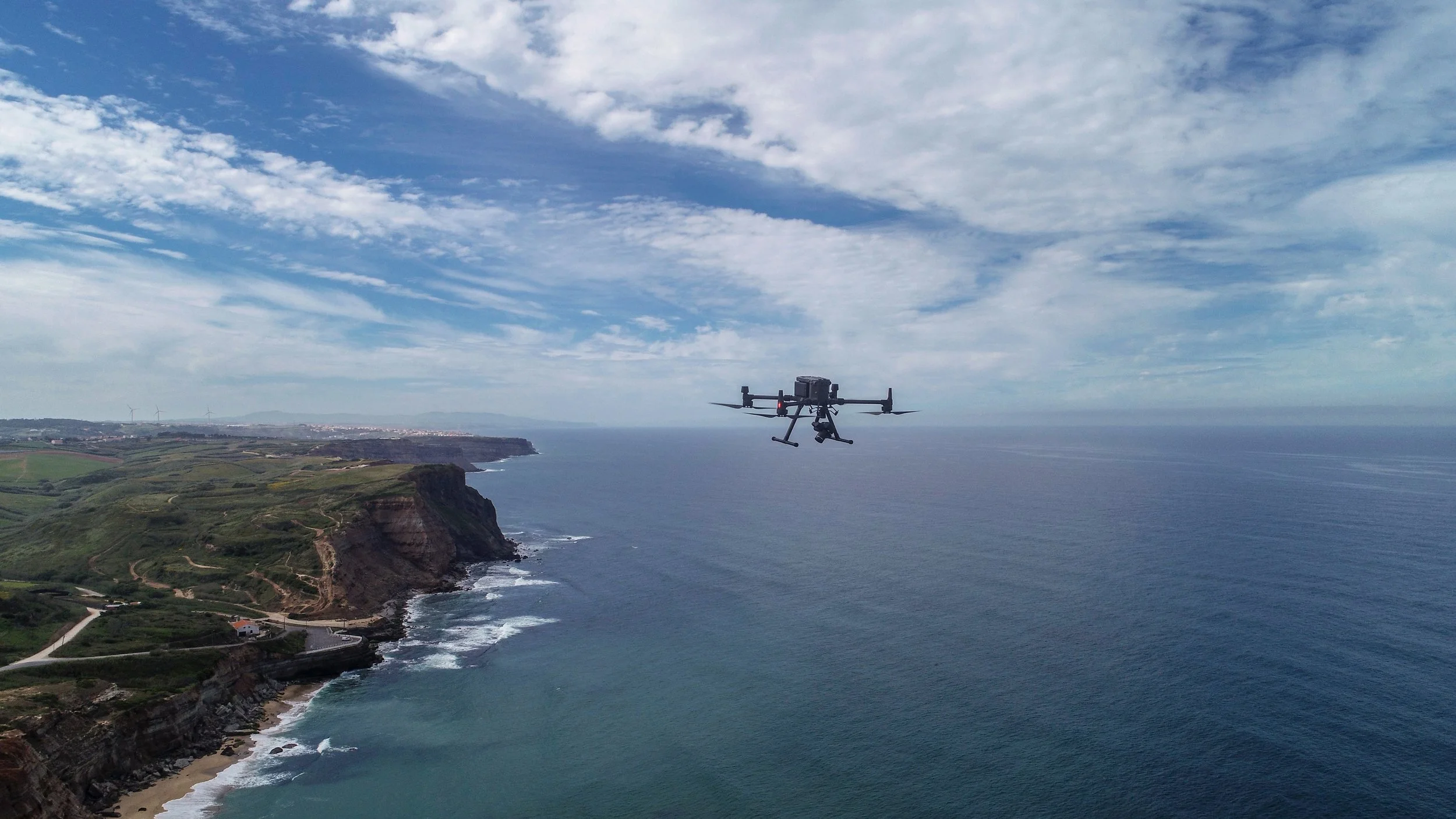 Drone voando acima de uma costa de arriba e mar, sob céu parcialmente nublado.