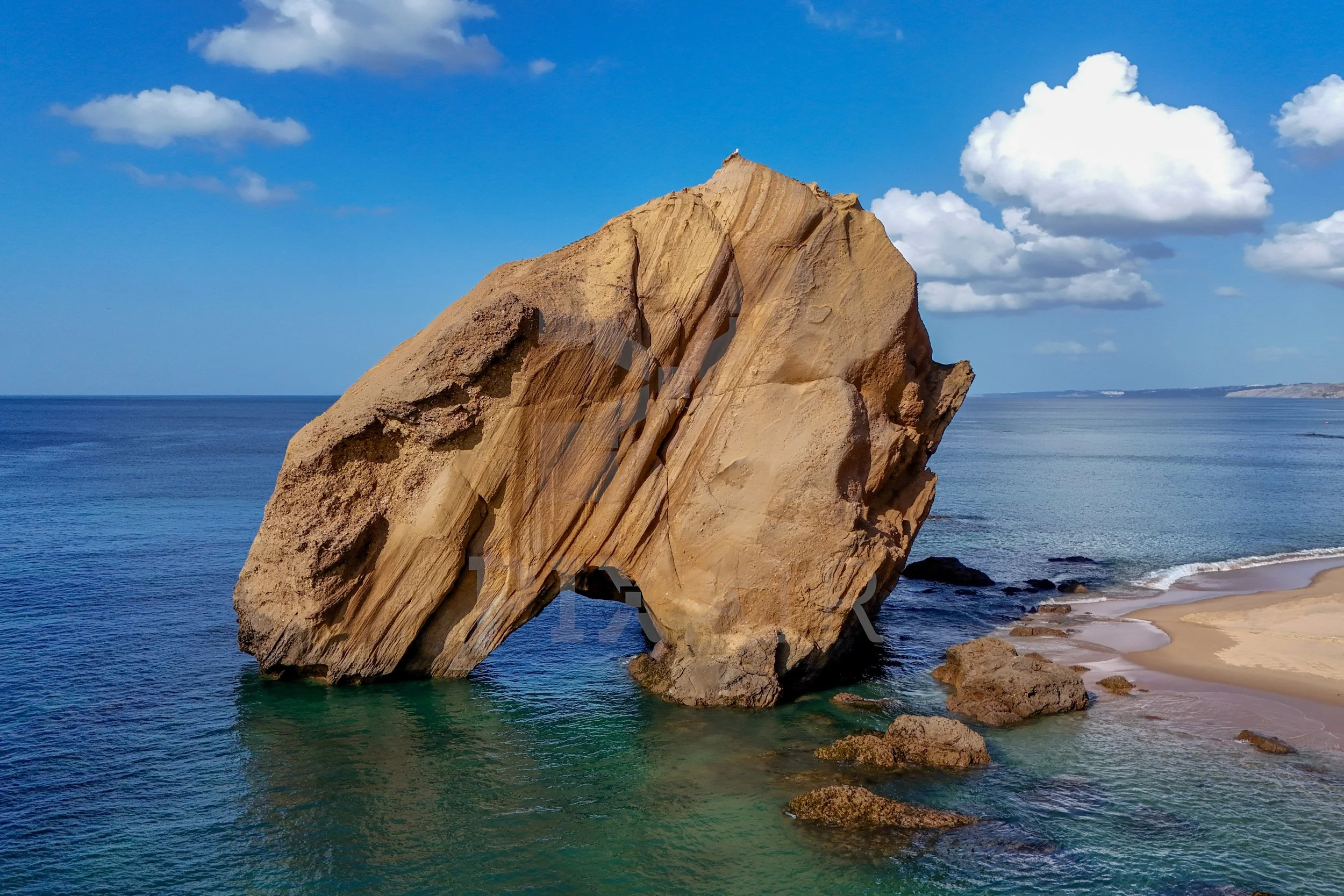 Grande rocha de formato irregular na orla do mar, com pequenas ondas ao redor e céu azul com algumas nuvens.