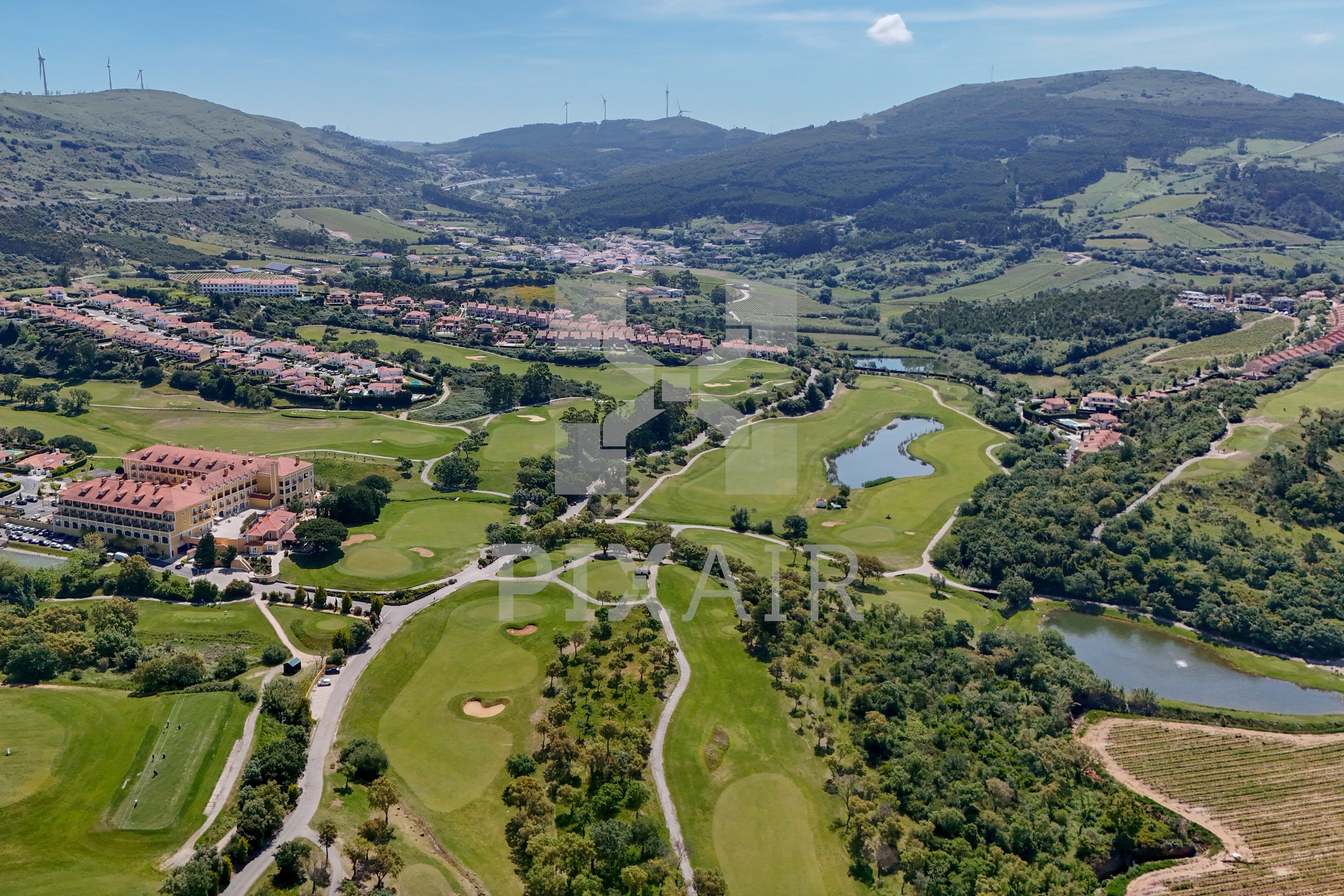 Vista aérea de um campo de golfe com rios, áreas verdes, árvores e edifícios ao redor, com colinas ao fundo.