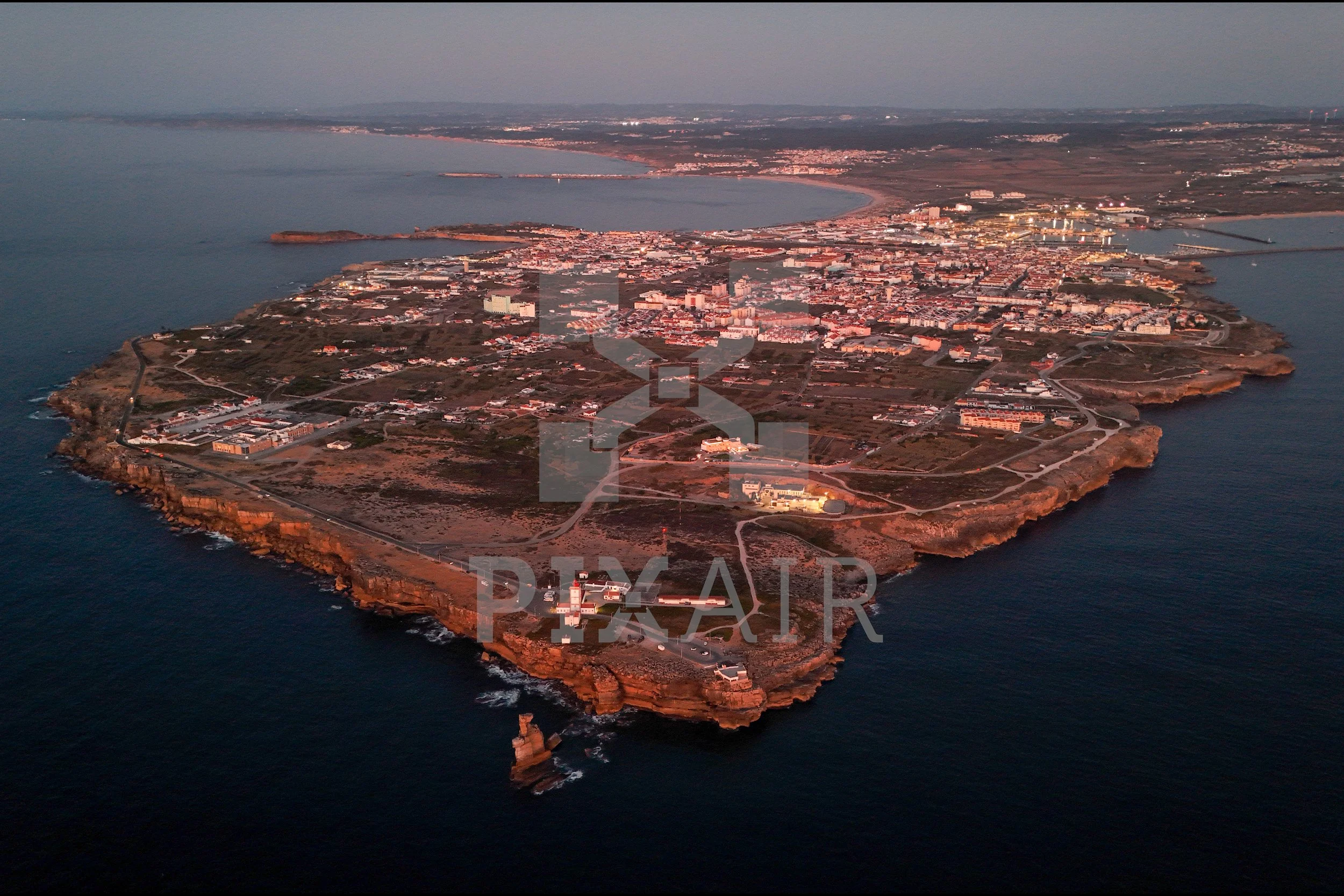 Ilha com costa rochosa e cidade ao fundo ao entardecer, vista aérea.