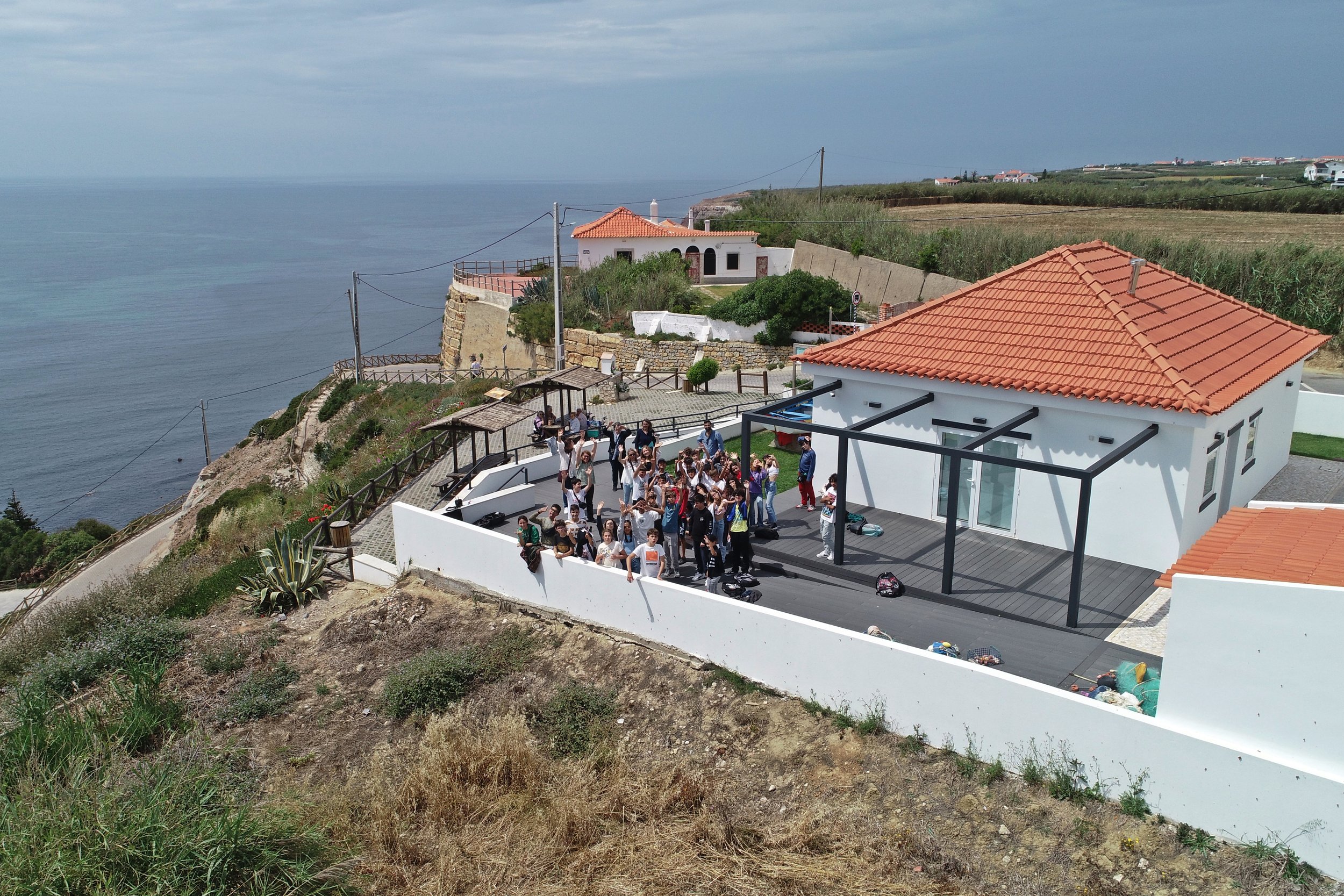 Grupo de pessoas, possivelmente estudantes, em uma área ao ar livre com piso de madeira, cercada por muros brancos, com uma casa branca de telhado de telhas vermelhas ao fundo, ao lado do mar.