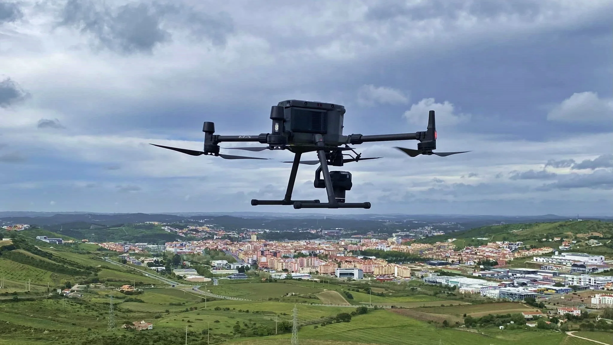 Drone sobrevoando uma cidade com áreas verdes e colinas sob céu nublado.