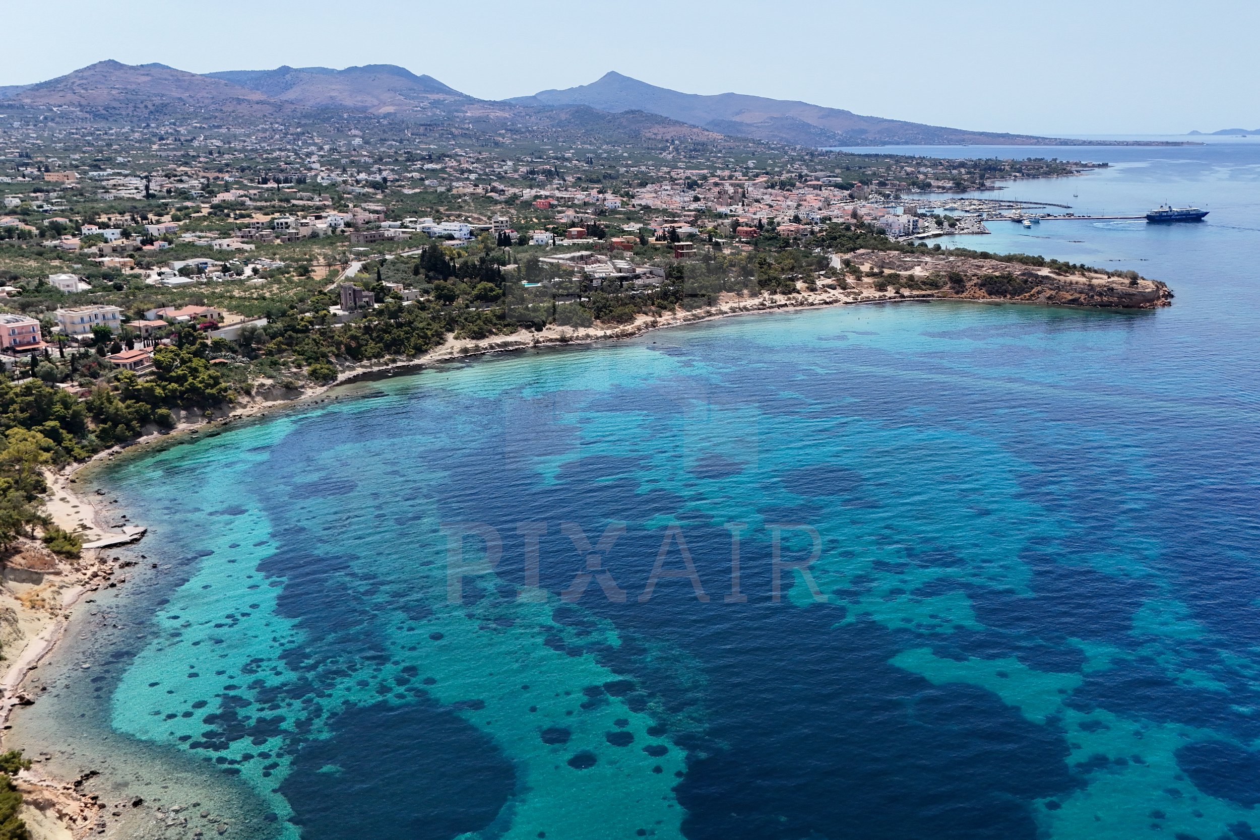 Vista aérea de uma baía com águas cristalinas em tons de azul e verde, cercada por um litoral rochoso, com uma cidade ao fundo e montanhas ao longe.