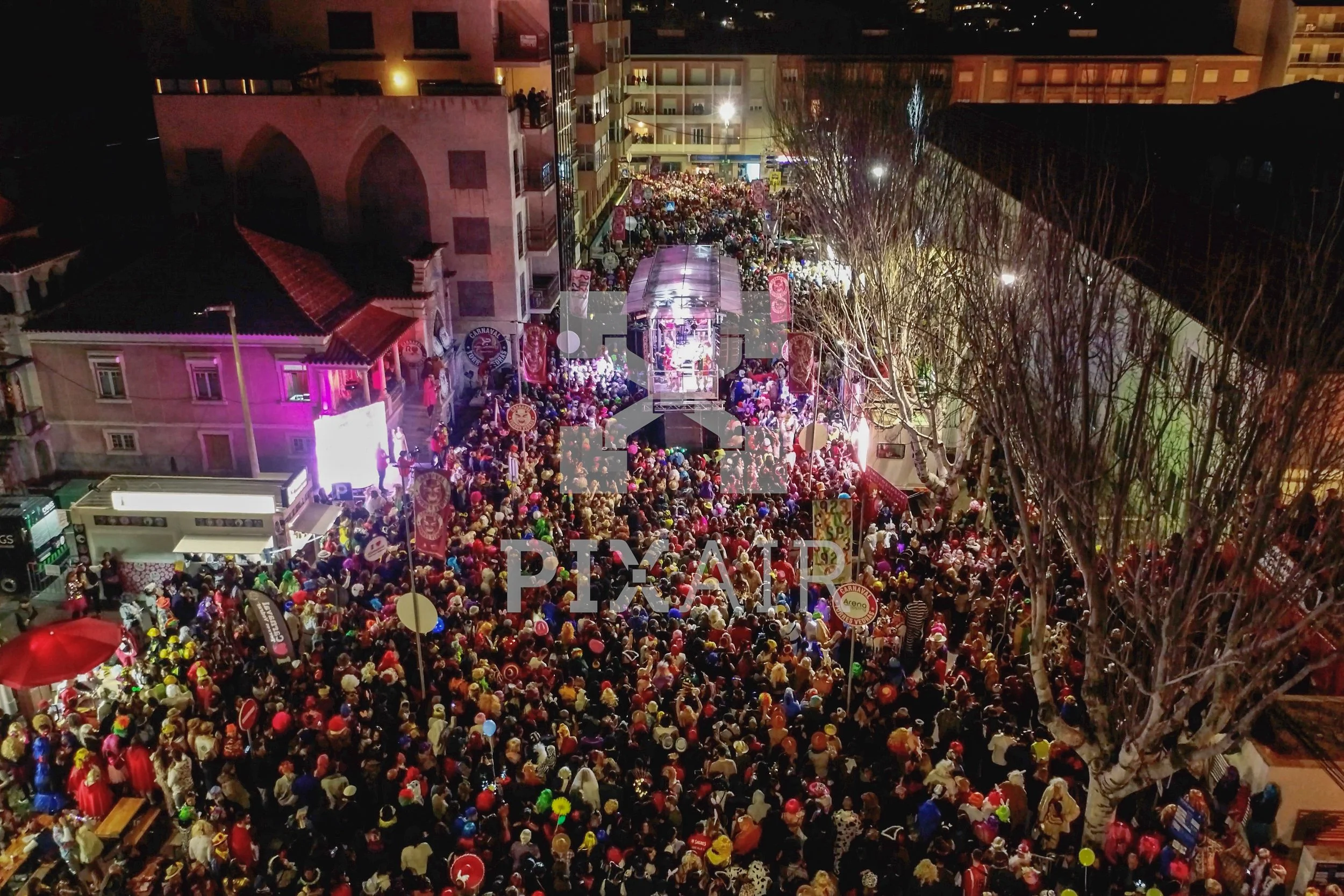 Grande aglomeração de pessoas em uma rua, com palco de show ao fundo, decorada para celebração noturna, com árvores desfolhadas na frente.