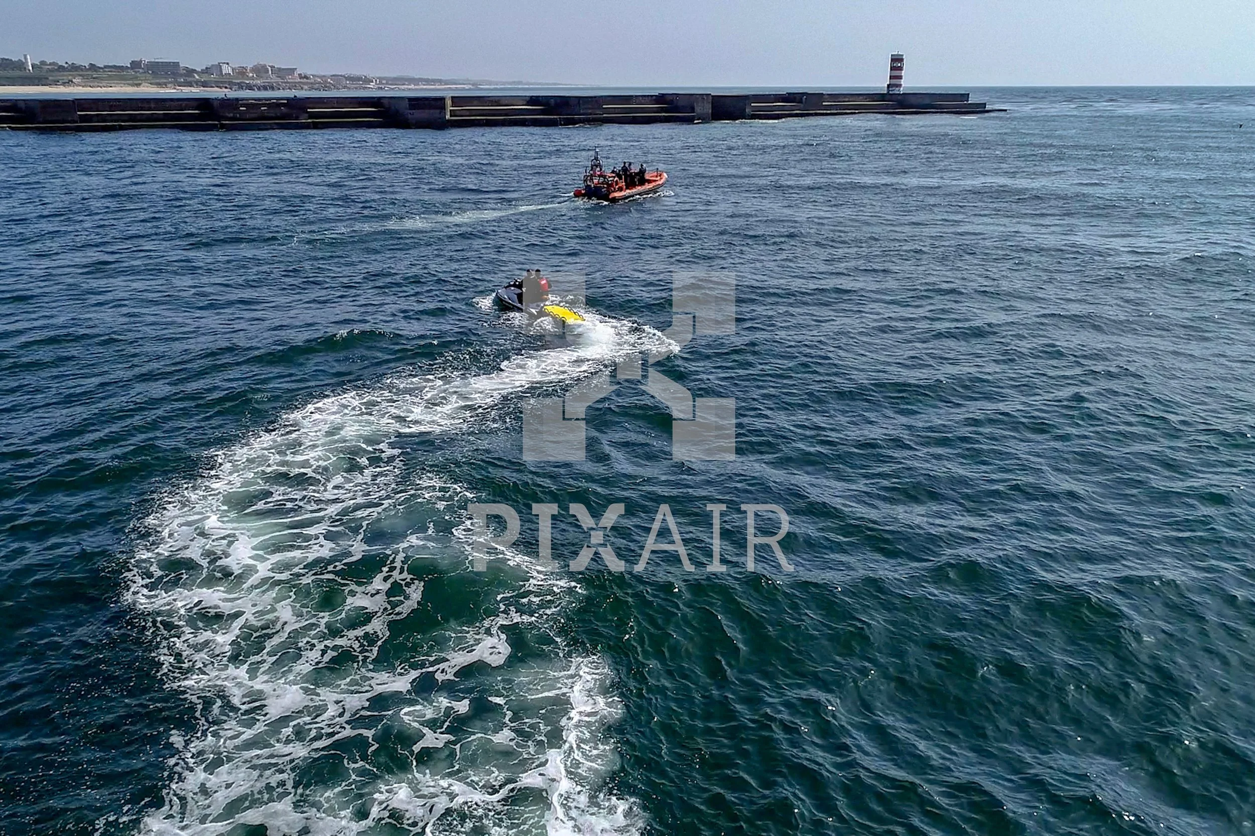 Imagem do mar com uma embarcação de resgate, possivelmente uma motoaquática, puxando uma bóia amarela. Ao fundo, há um píer com um farol na extremidade e uma cidade na linha do horizonte.