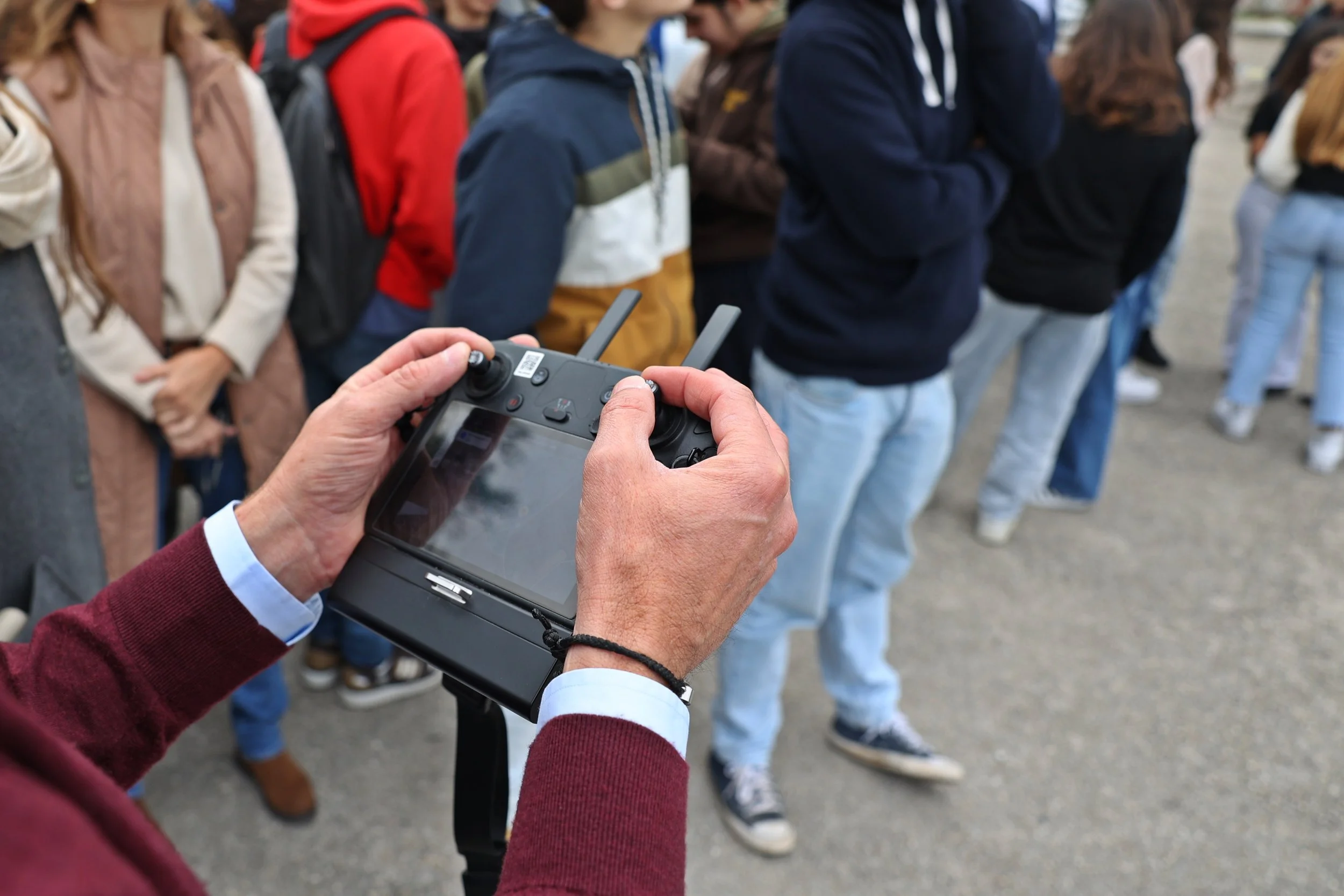 Pessoa segurando controle remoto de drone enquanto observa uma fila de pessoas ao ar livre.