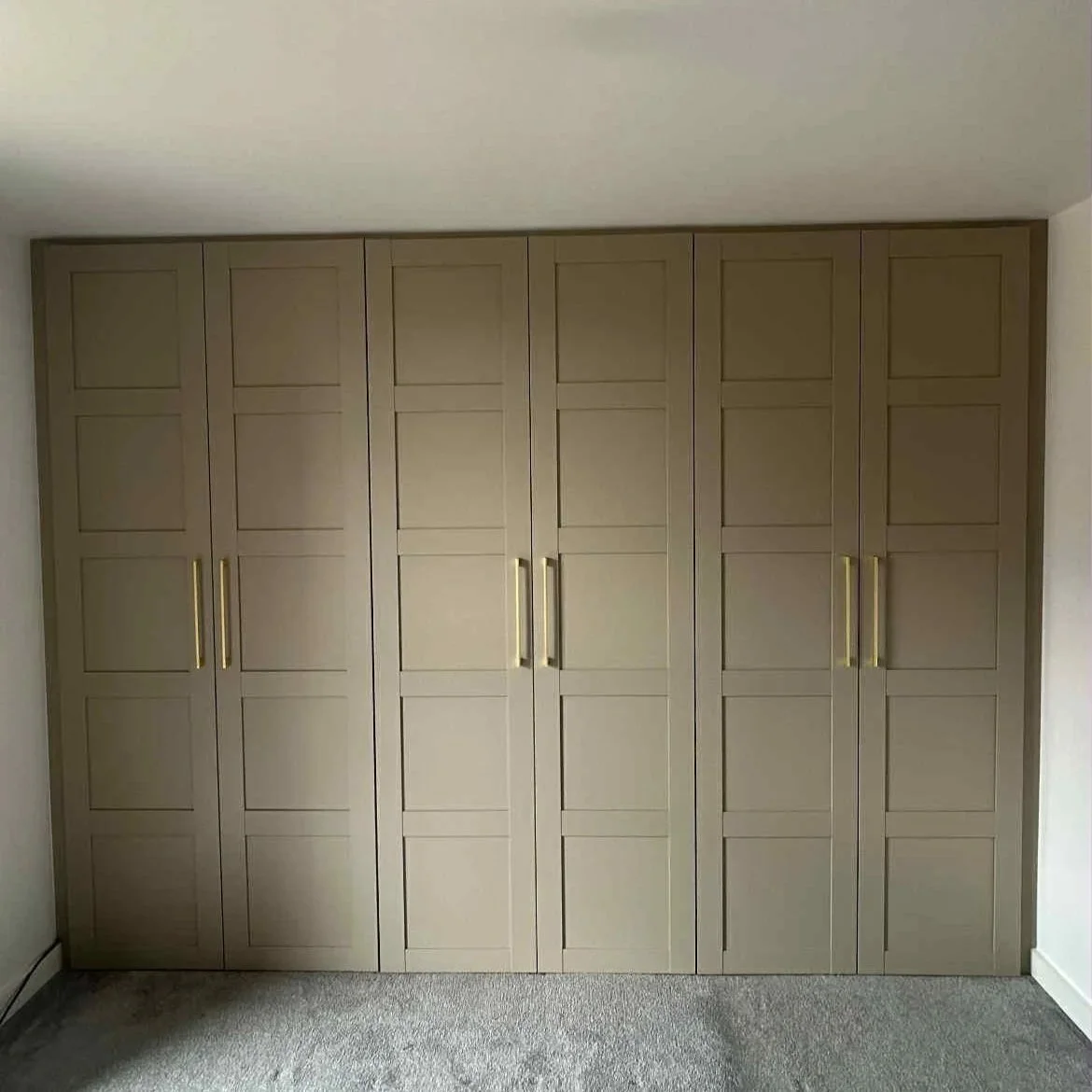 A beige, paneled closet with five doors and gold handles in a room with light-colored walls and carpeted floor.
