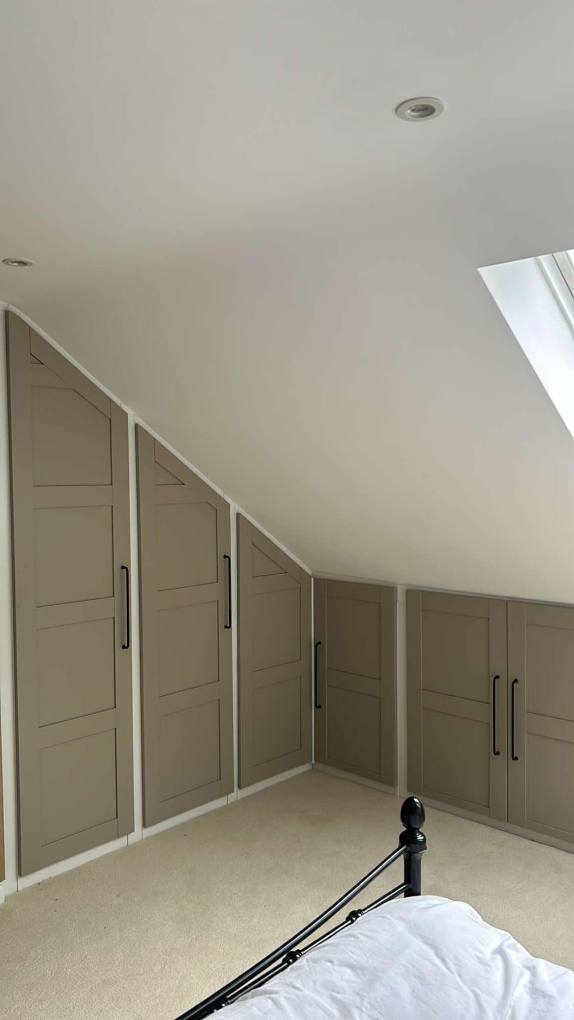 A bedroom with beige built-in corner cabinets with black handles under a sloped ceiling with a small skylight window.