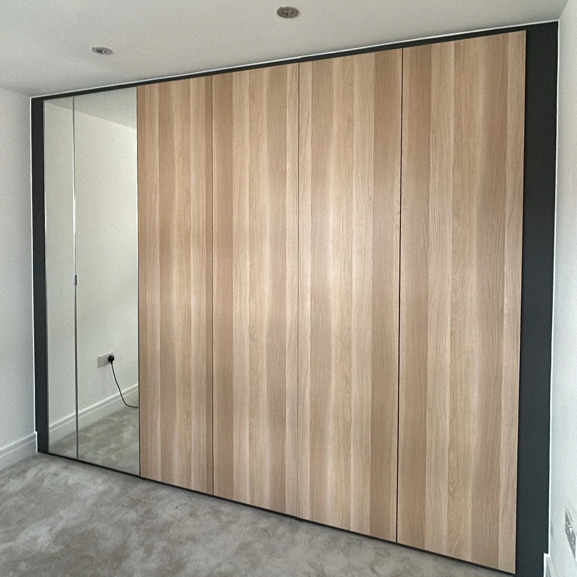 A modern built-in wardrobe with light wood panels and a mirror on the left side, set in a room with white walls and beige carpet.