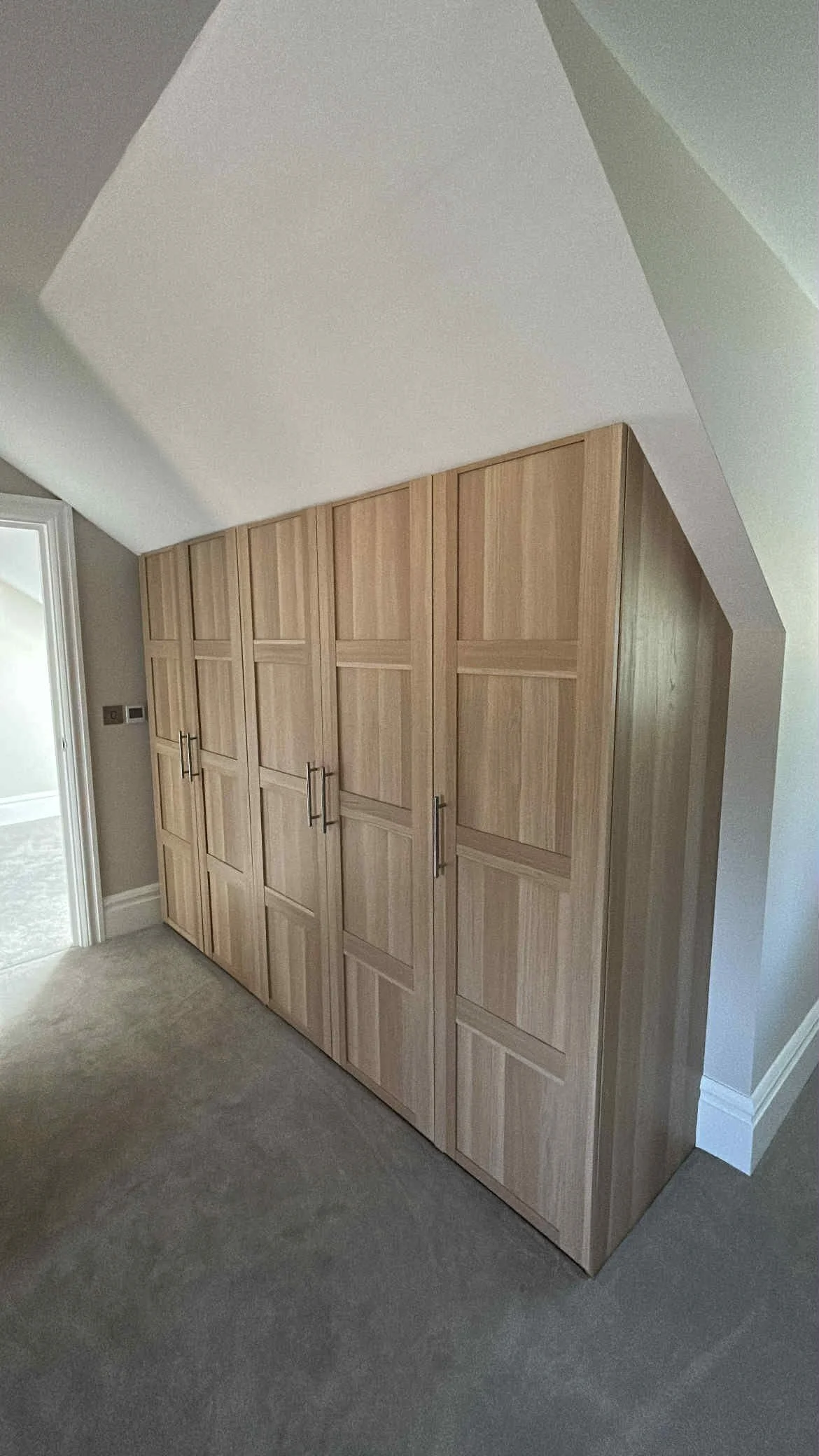 A large wooden closet with multiple doors and metal handles, placed in a corner of a room with beige carpet and light-colored walls.