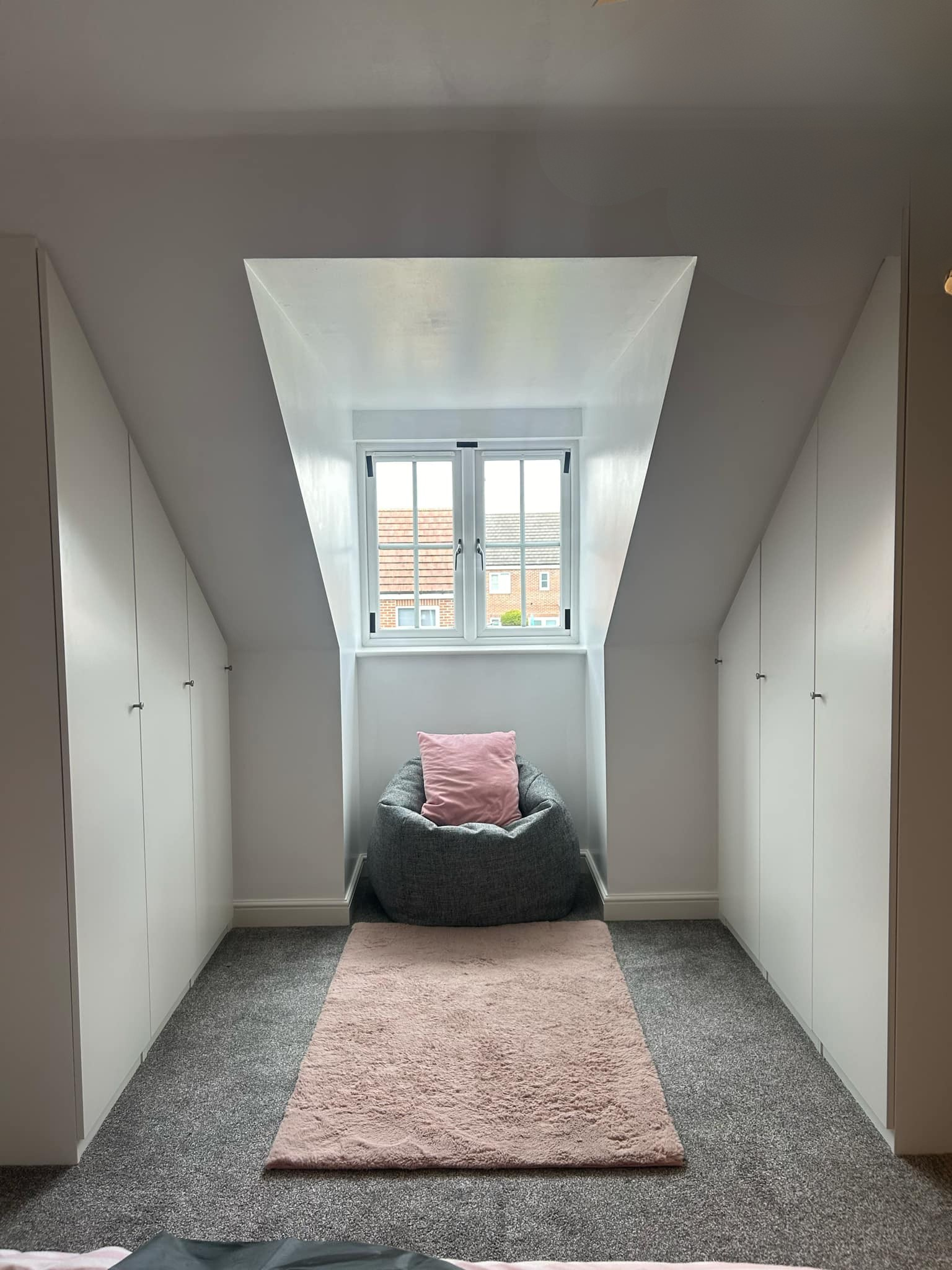 A cozy attic room with white built-in cabinets on both sides, a pink rug on gray carpet, a gray bean bag chair with a pink pillow, and a window showing neighboring buildings.