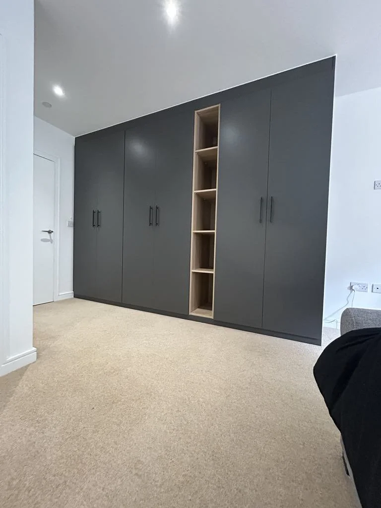 A large dark gray wardrobe with multiple doors and black handles, and an open wooden shelf in the middle, located in a room with beige carpet and white walls.