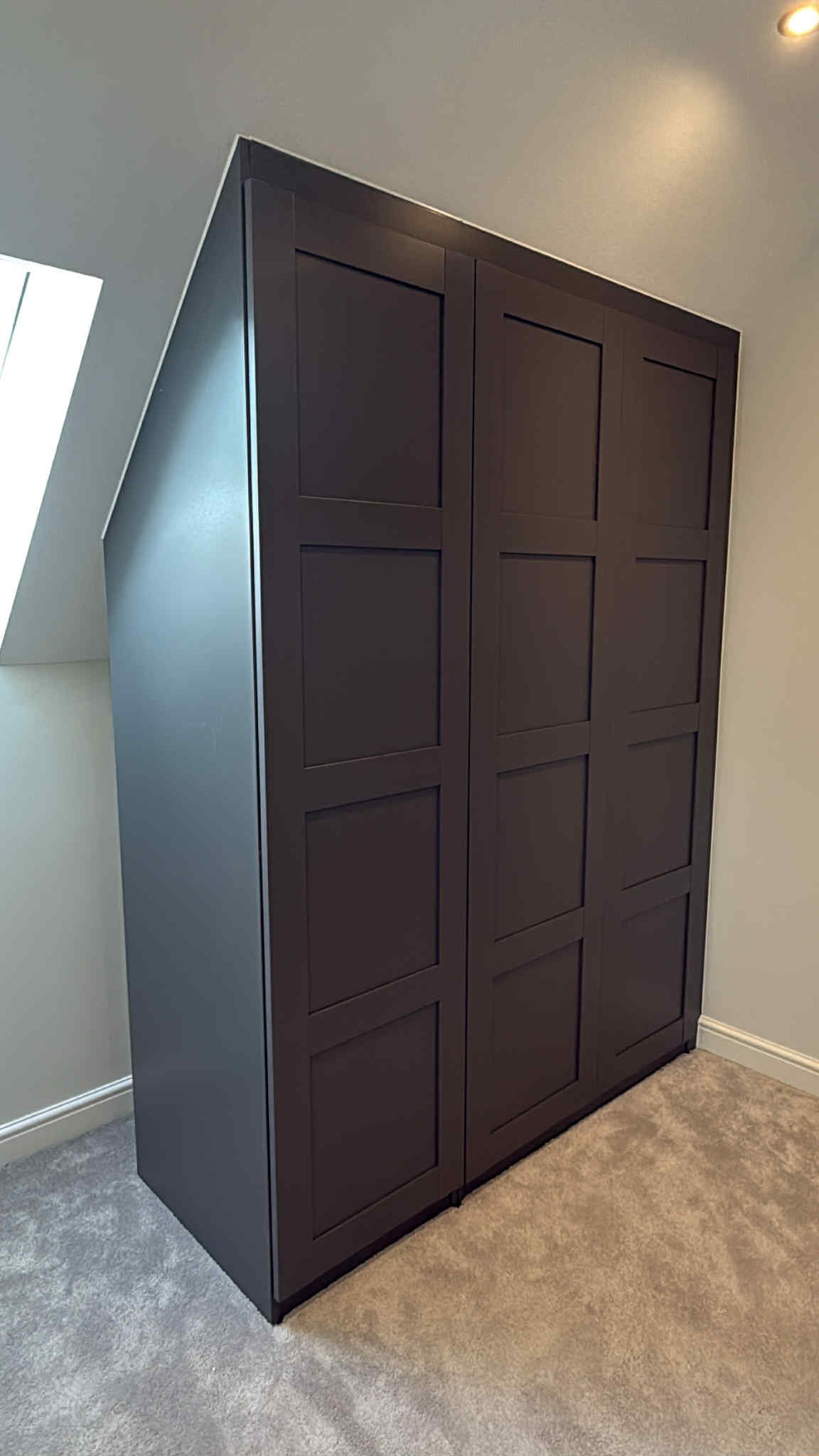 Tall black wooden wardrobe with a grid-panelled door design in a room next to a window sloped ceiling.