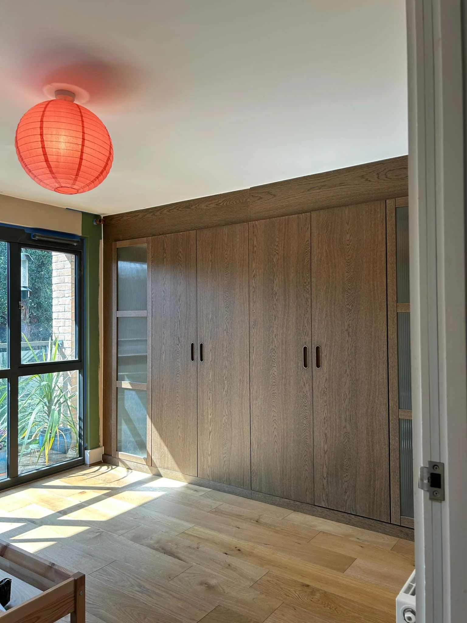 A room with a wooden wardrobe, a large glass door, a red paper lantern hanging from the ceiling, and sunlight streaming through the door onto the wooden floor.