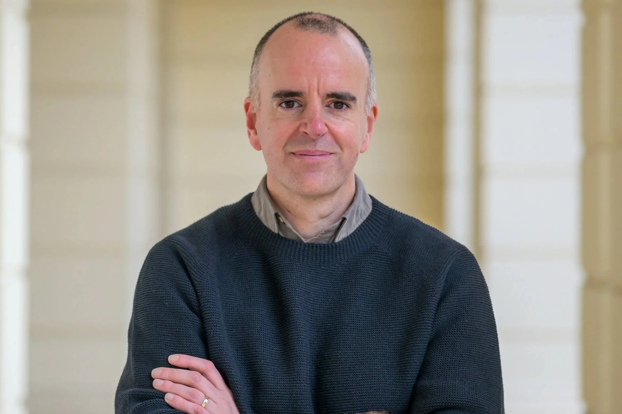 A man with short gray hair and a light complexion is standing with his arms crossed, wearing a dark sweater over a collared shirt, in front of a blurred indoor background.
