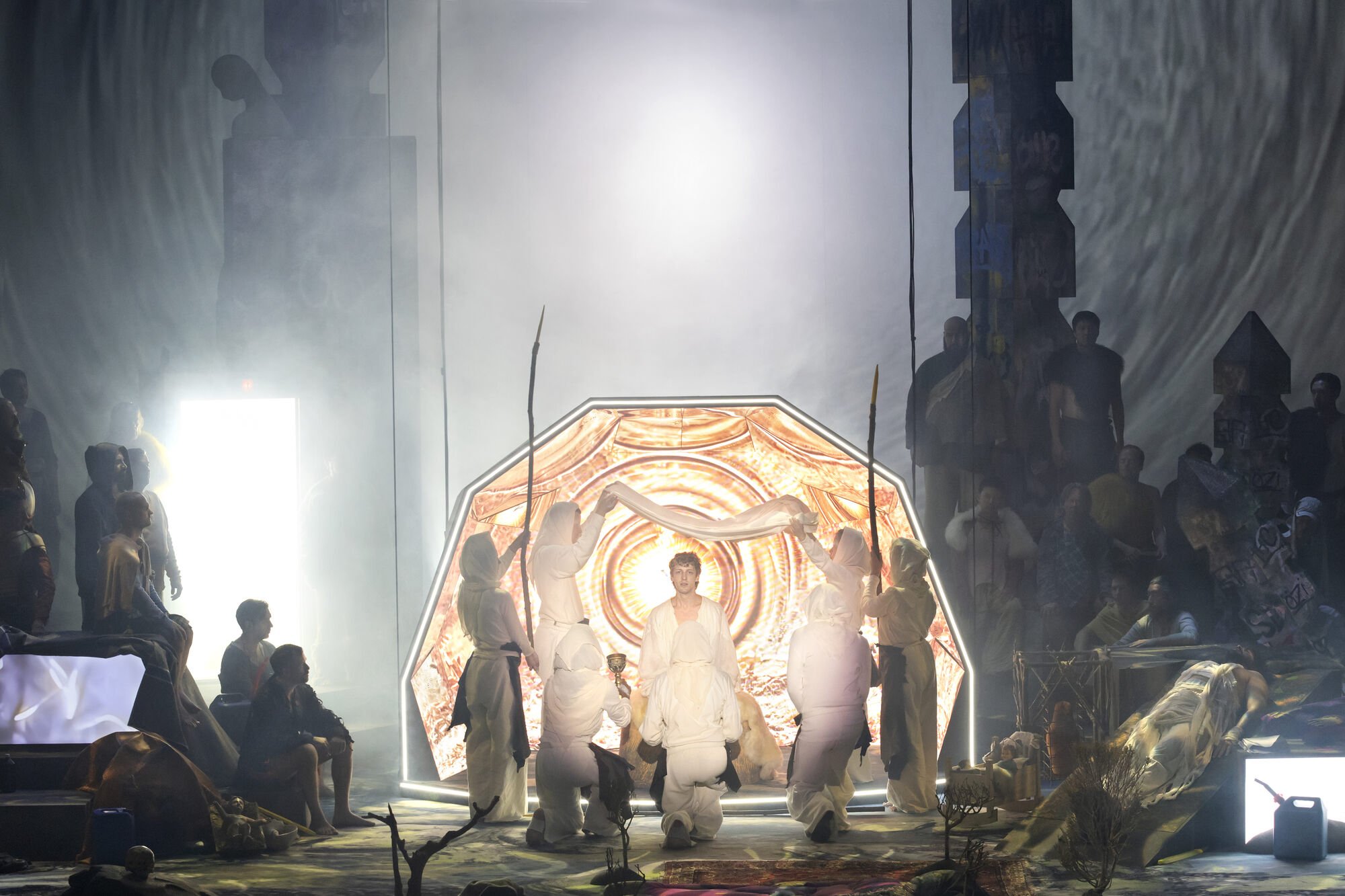 A theatrical scene with performers dressed in white, surrounded by dramatic lighting and fog, with an illuminated backdrop resembling an eye.