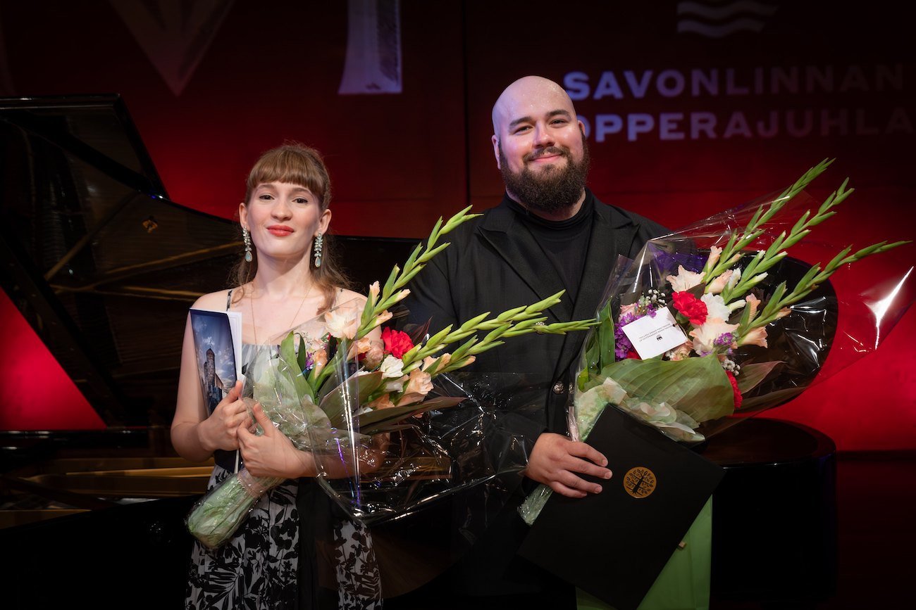 Elsa Angervo and Martin Iivarinen standing in front of a red background, each holding a bouquet of flowers, with a black grand piano behind them.