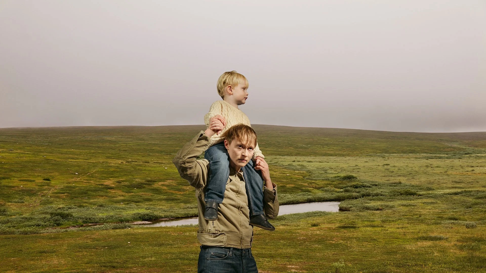 A man carrying a young girl on his shoulders through an open grassy landscape with hills in the distance under a cloudy sky.