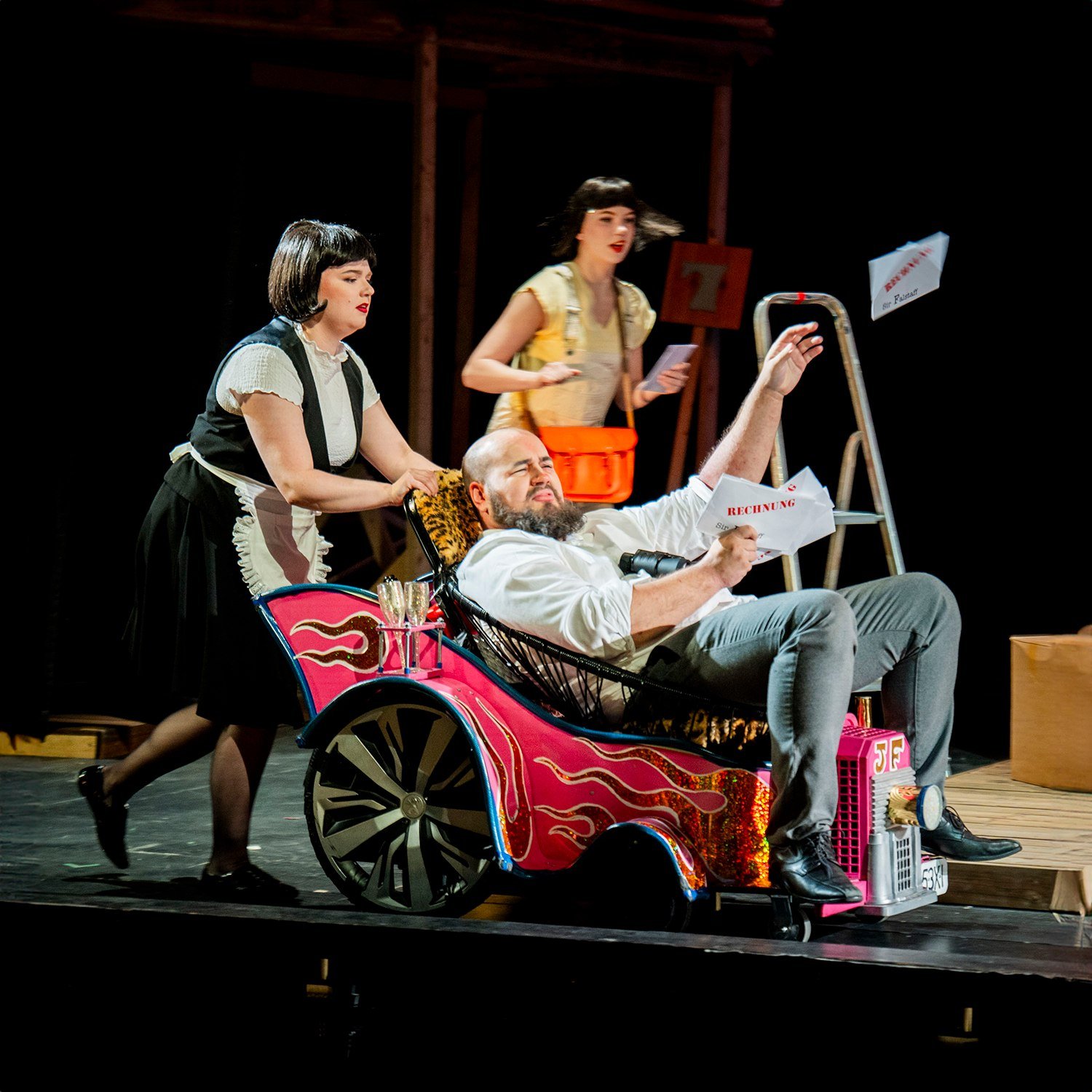 Three actors on stage in a theatrical play. One actor is sitting in a small, colorful car prop, holding a document, with an exaggerated facial expression. Another actress is pushing the car, and a third actress is standing behind, holding a piece of paper. The scene appears comedic with props around them, including a ladder and signs.