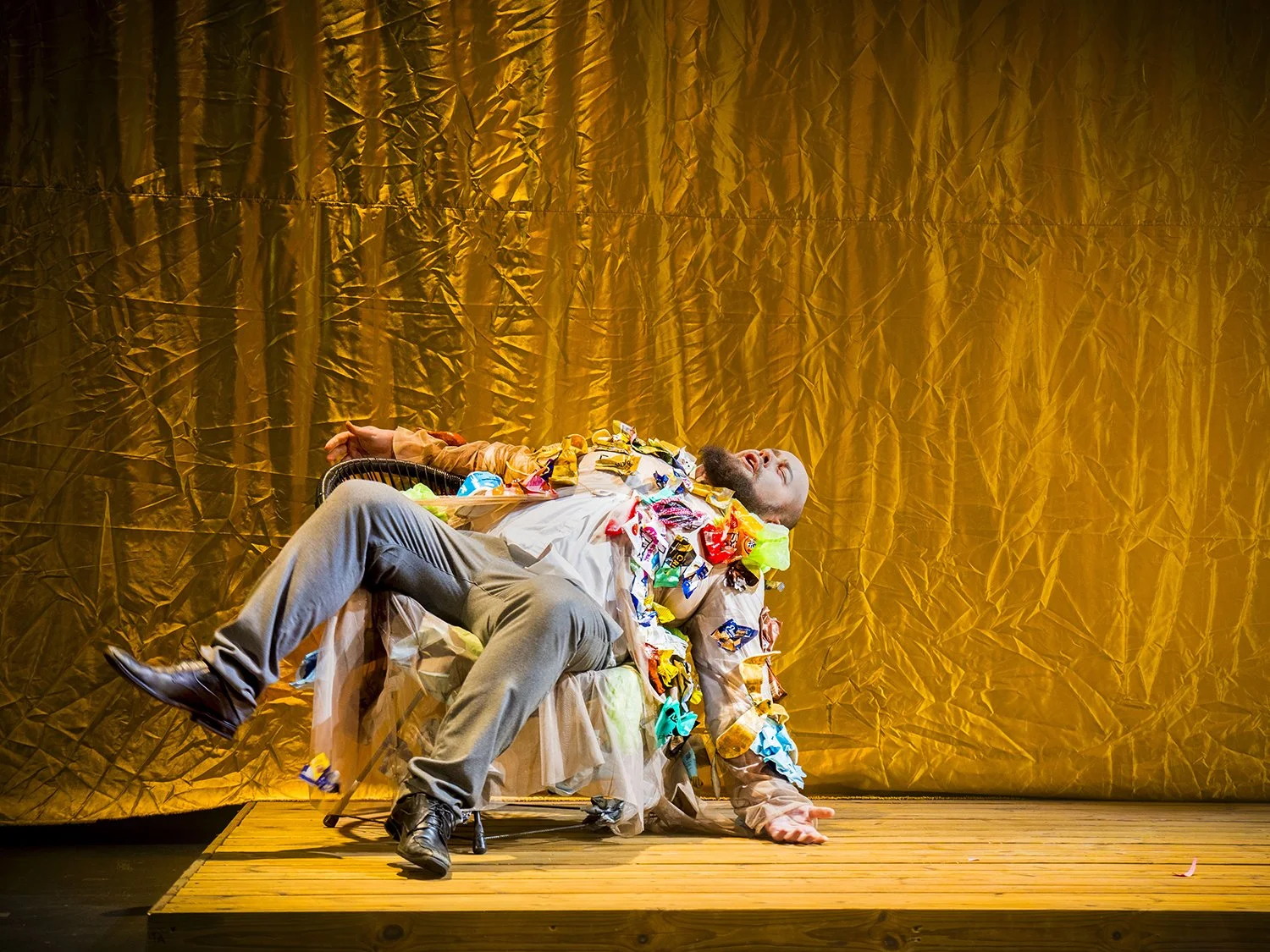Martin Iivarnen in-costume as Sir John Falstaff in the opera Die lustigen Weiber von Windsor, lying on a chair on a wooden stage with a crumpled gold backdrop, wearing a shirt covered in colorful candy wrappers.