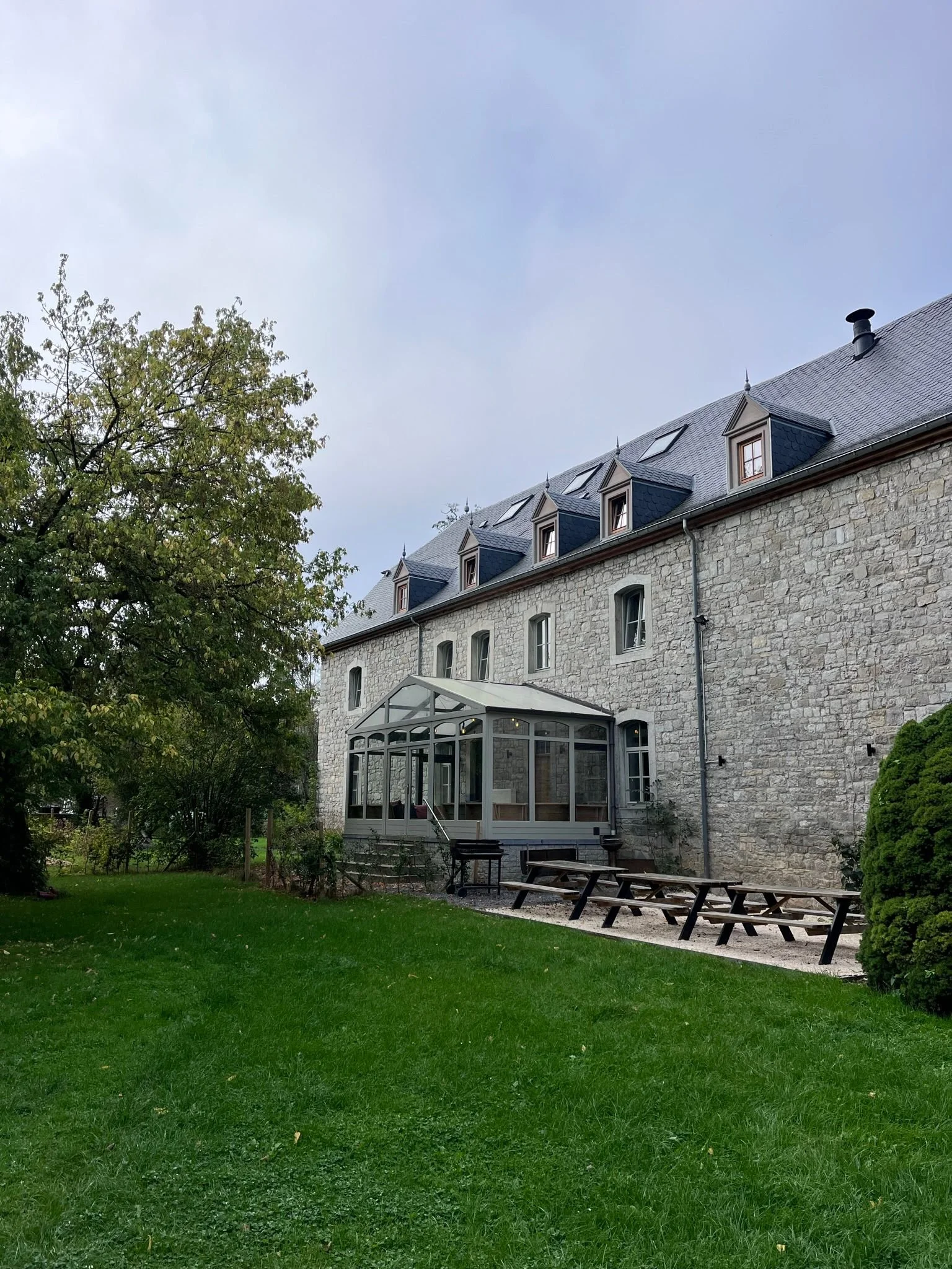 Grote stenen woning met een glazen veranda en een tuin met gras, bomen en picknicktafels.