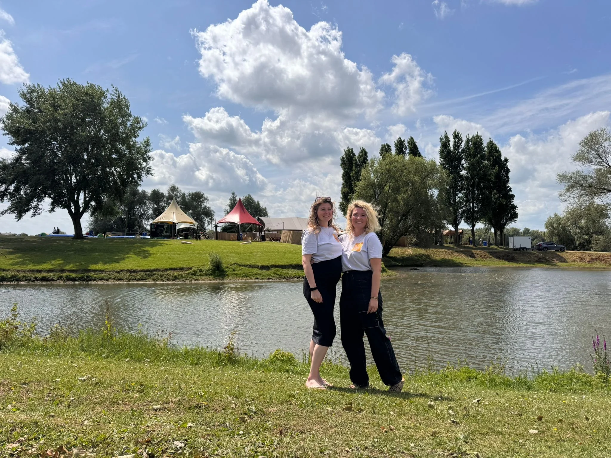 Twee vrouwen staan samen aan de rand van een rivier, glimlachend naar de camera, met een groene oever en tenten op de achtergrond onder een deels bewolkte blauwe hemel.