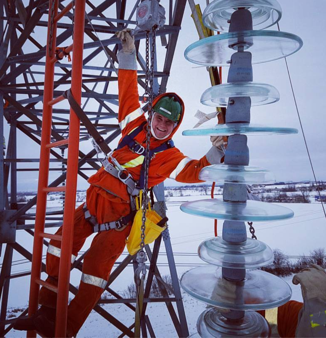 A utility worker in safety gear working on electrical equipment on a tall structure during winter, with snow-covered ground in the background.