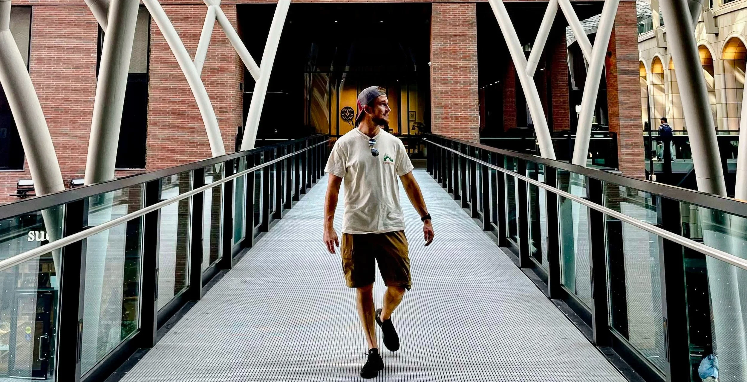 A man wearing a baseball cap, t-shirt, and shorts walking on a glass enclosed bridge inside a modern building with brick walls and large windows.