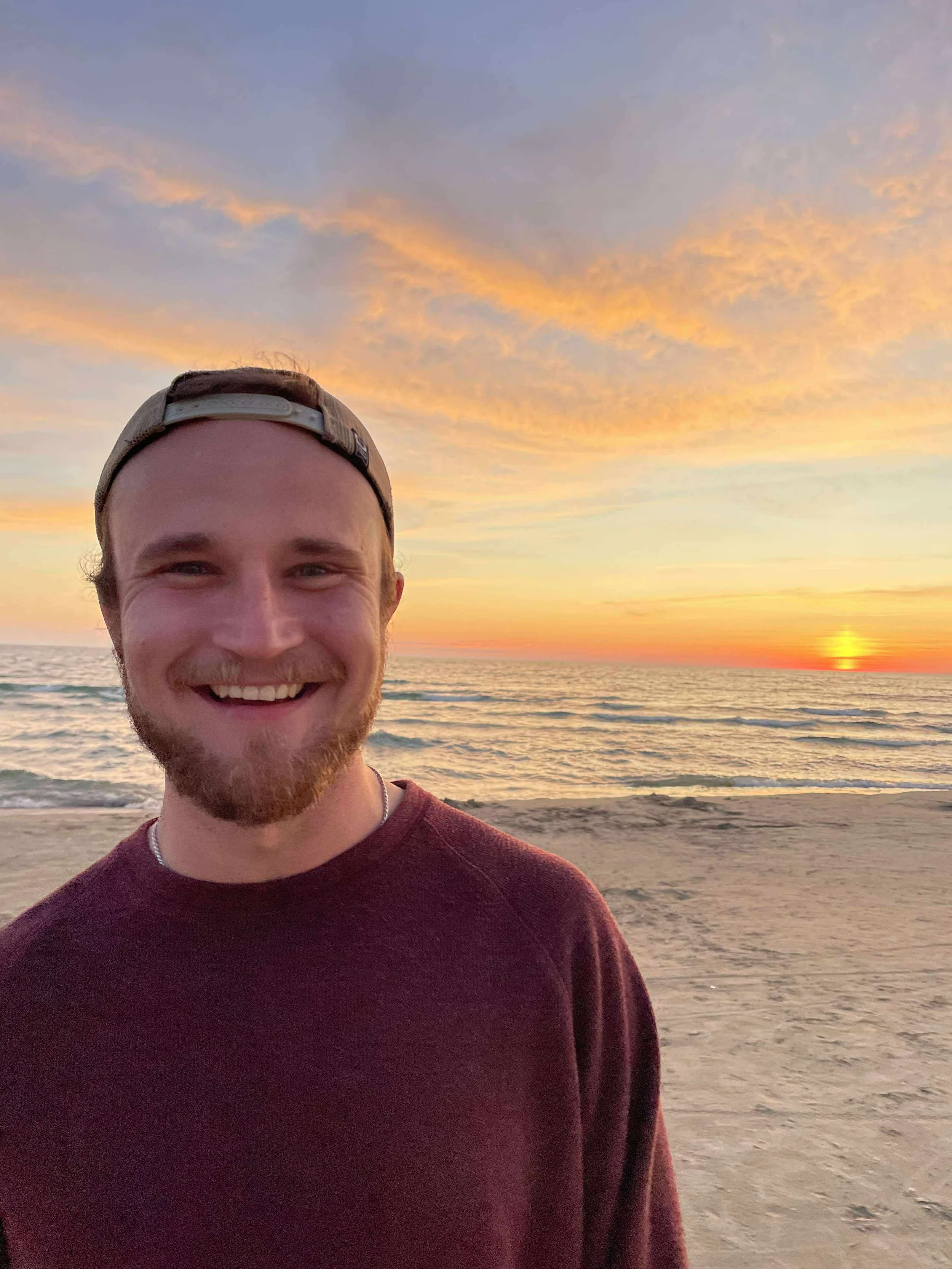 A smiling man with a beard and baseball cap standing on the beach at sunset, with the ocean, sandy shore, and colorful sky in the background.
