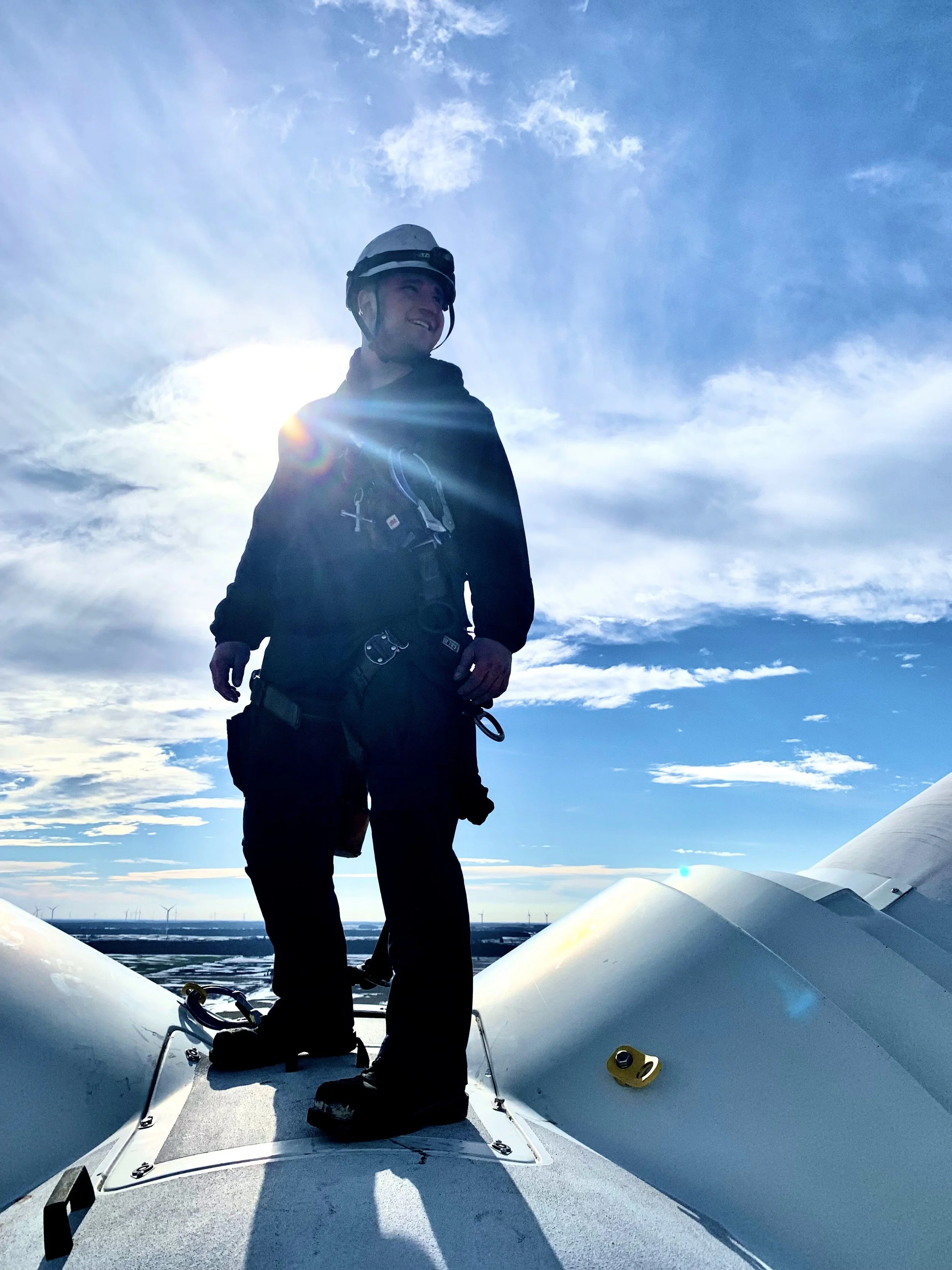 A person with a helmet and outdoor gear standing on the deck of a boat with the sky and clouds in the background, and water and wind turbines visible in the distance.