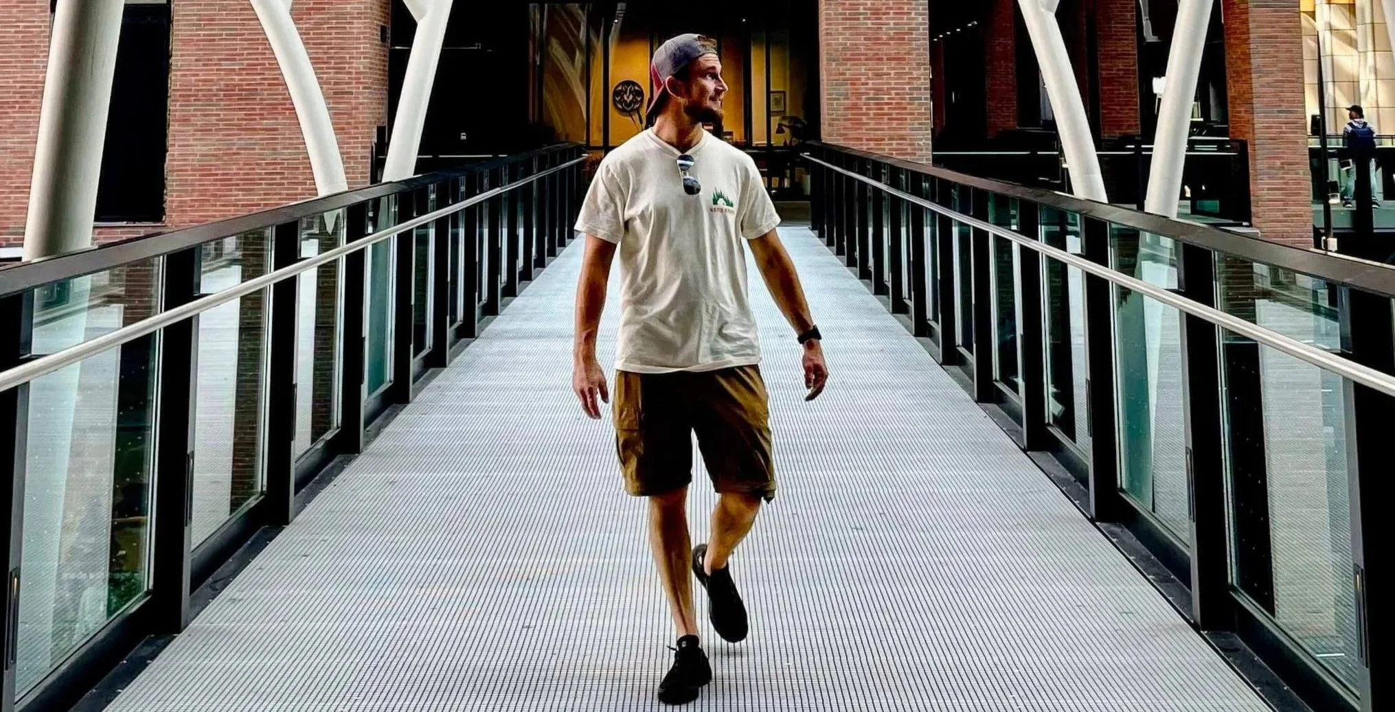 A man walking on a glass-walled pedestrian bridge inside a modern building with brick walls, wearing a white t-shirt, shorts, a backward cap, and sunglasses hanging from his shirt.
