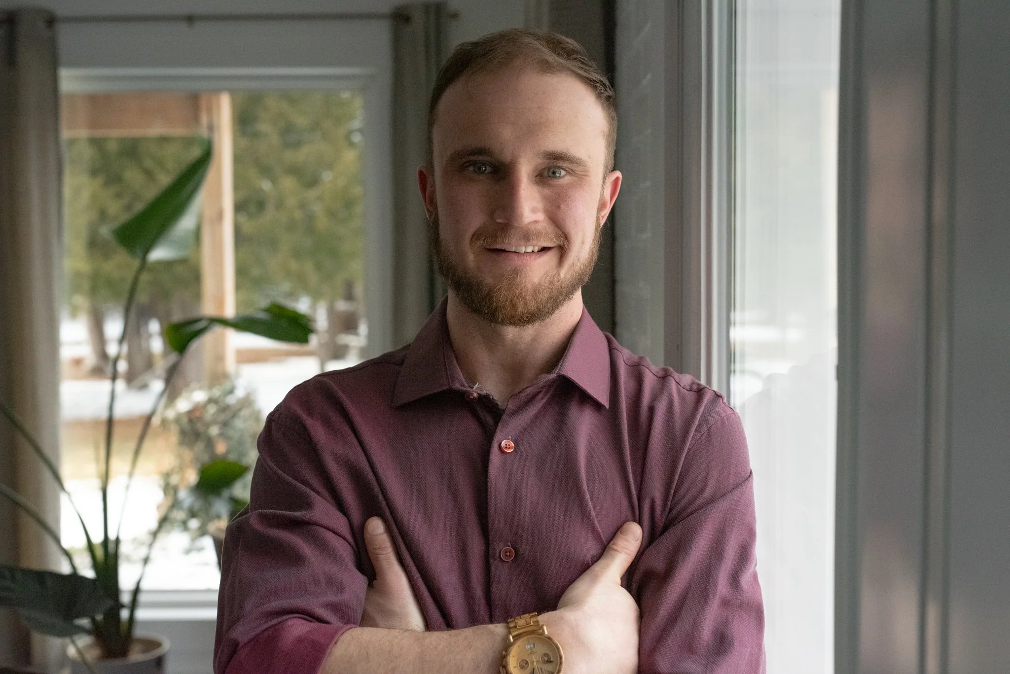 A young man with a beard and short hair standing indoors near a window, crossing his arms, wearing a maroon shirt and a gold watch.