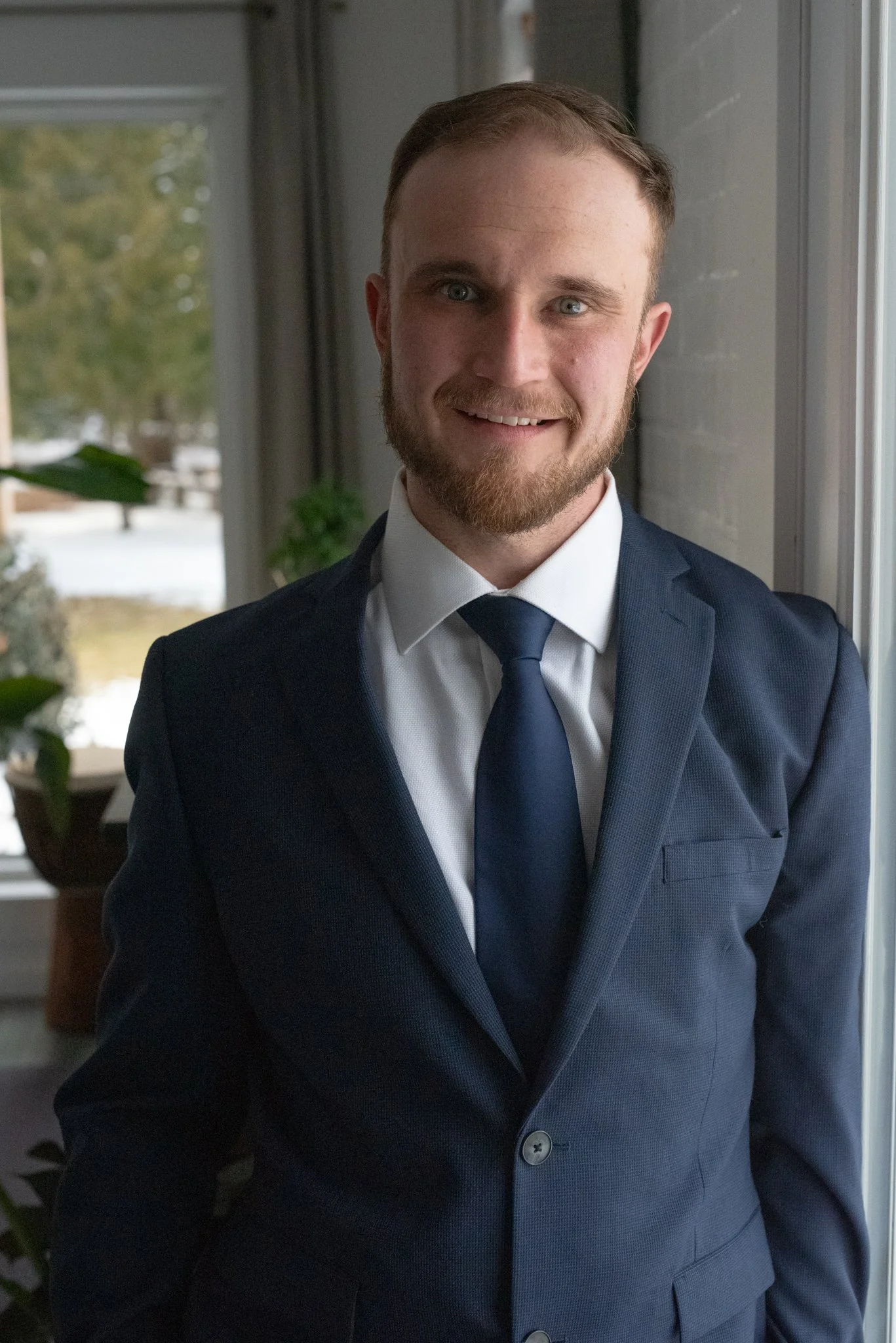 A young man with light skin, brown hair, and a beard, dressed in a navy blue suit, white shirt, and navy tie, standing near a window with a blurred indoor background that includes a plant and a snowy outdoor scene.