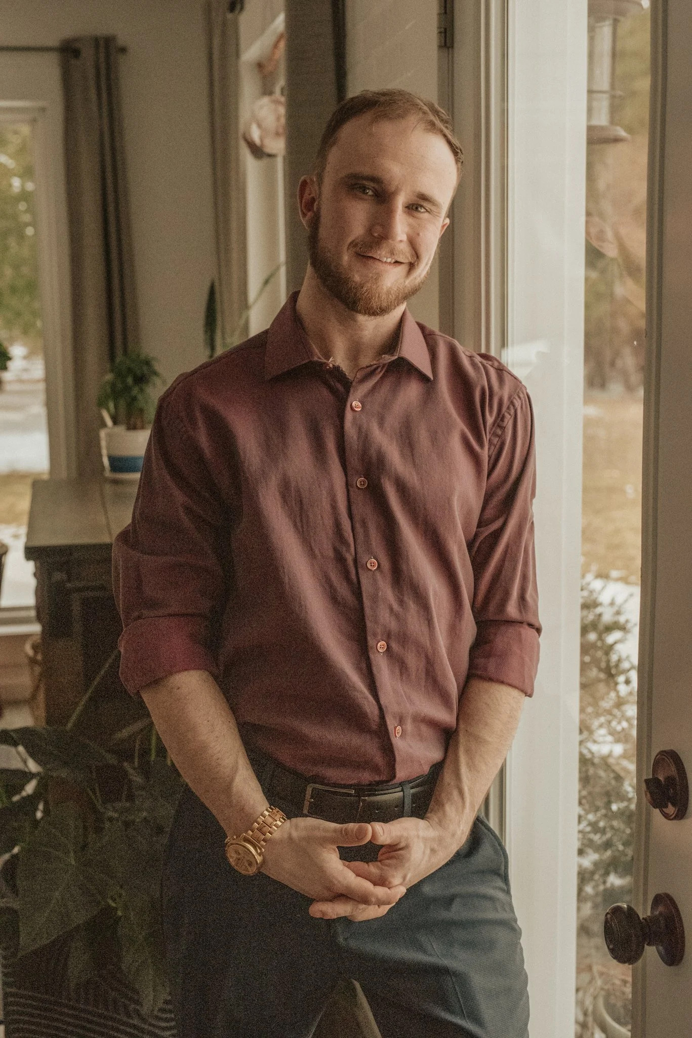 A young man with light skin, short hair, and a beard smiling, standing indoors near a window with a view of trees and winter scenery. He is wearing a maroon button-up shirt with the sleeves rolled up and a gold watch on his left wrist.