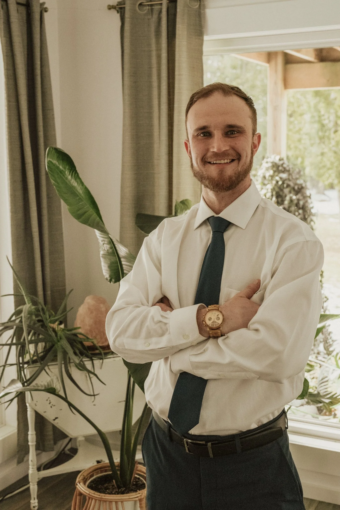 A man wearing a white shirt, dark tie, with his arms crossed, smiling in a room with plants and a window.