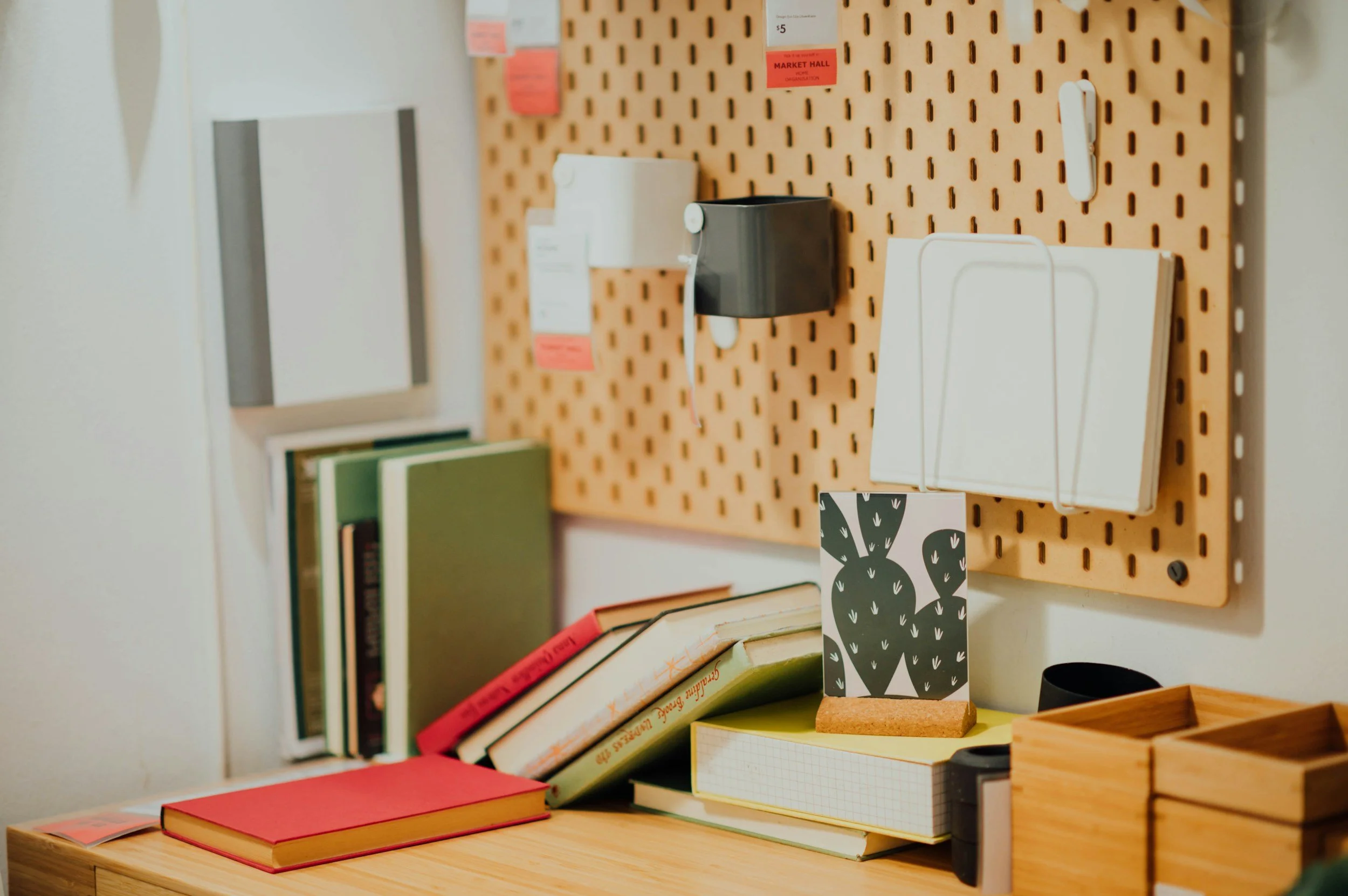 A workspace with a light wood desk, books, notebooks, and a pegboard with various office supplies and decor. There are shelves with books, a small black speaker, and a decorative card with a cactus illustration.