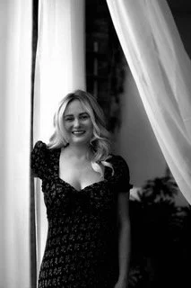A smiling woman with wavy blonde hair standing near curtains by a window in a black dress.