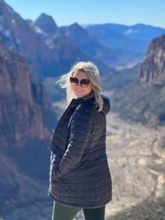 A woman with blonde hair wearing sunglasses and a dark jacket smiling outdoors with a mountainous landscape in the background.