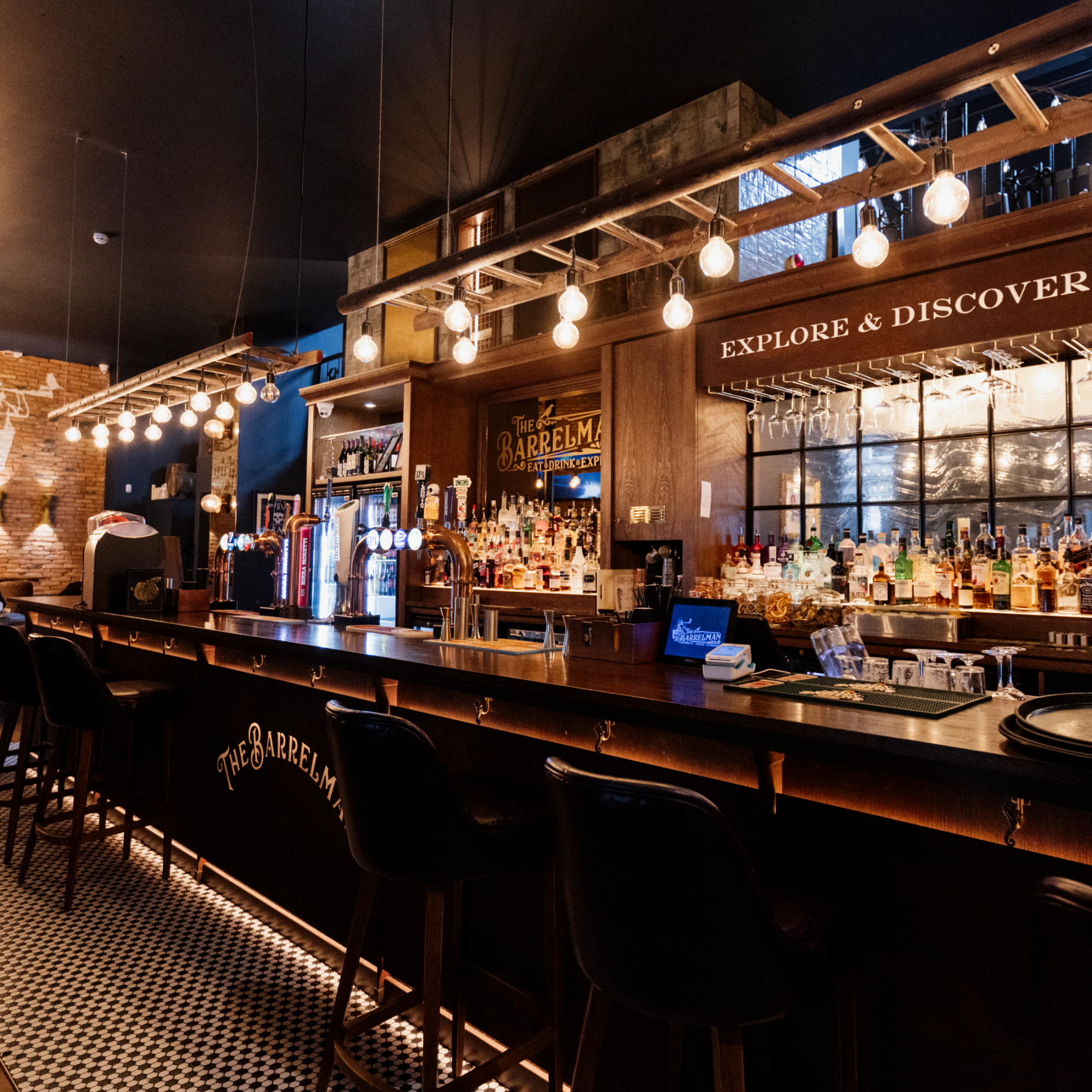 Interior of a bar with hanging light bulbs, a wooden bar counter, and shelves with liquour bottles behind it, with a sign that reads 'Explore & Discover'.