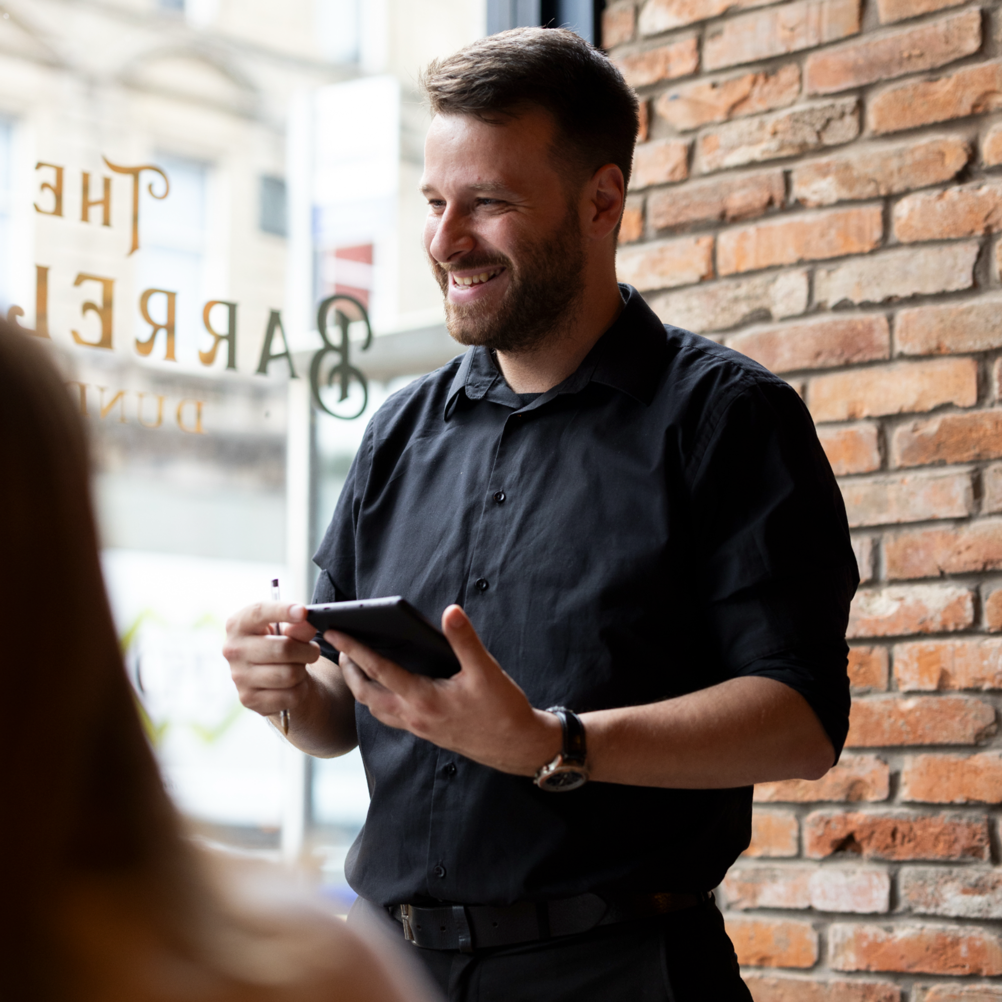 A man with dark hair, a beard, wearing a black shirt, smiling and holding a device, standing in front of a brick wall inside a building.