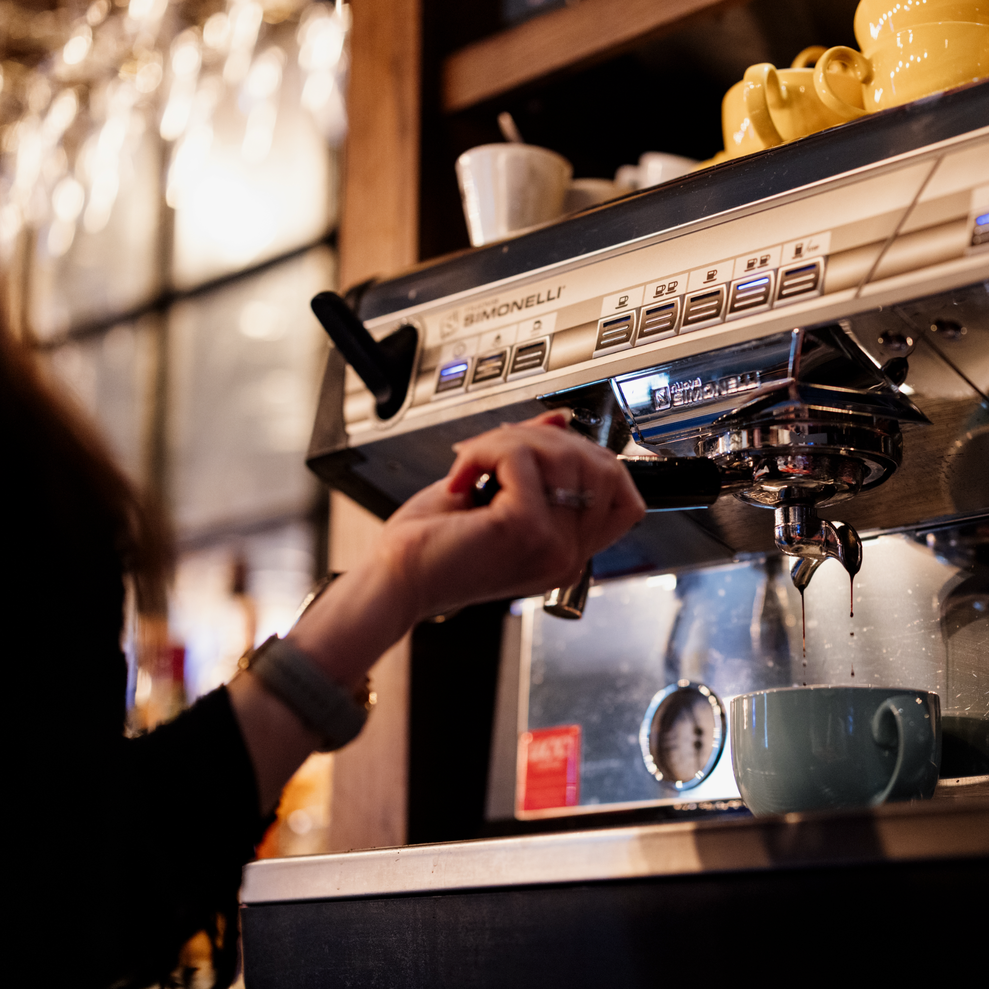 Close-up of a coffee machine with a row of yellow cups and a person's hand pressing buttons on the machine.