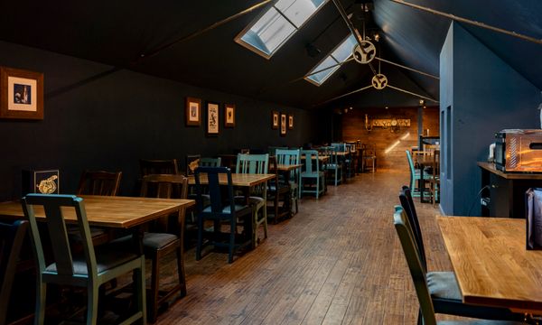 Interior of a cozy restaurant with wooden floors, blue and dark walls, framed pictures, and a variety of wooden tables and chairs, some painted blue, arranged along the sloped ceiling with skylights.