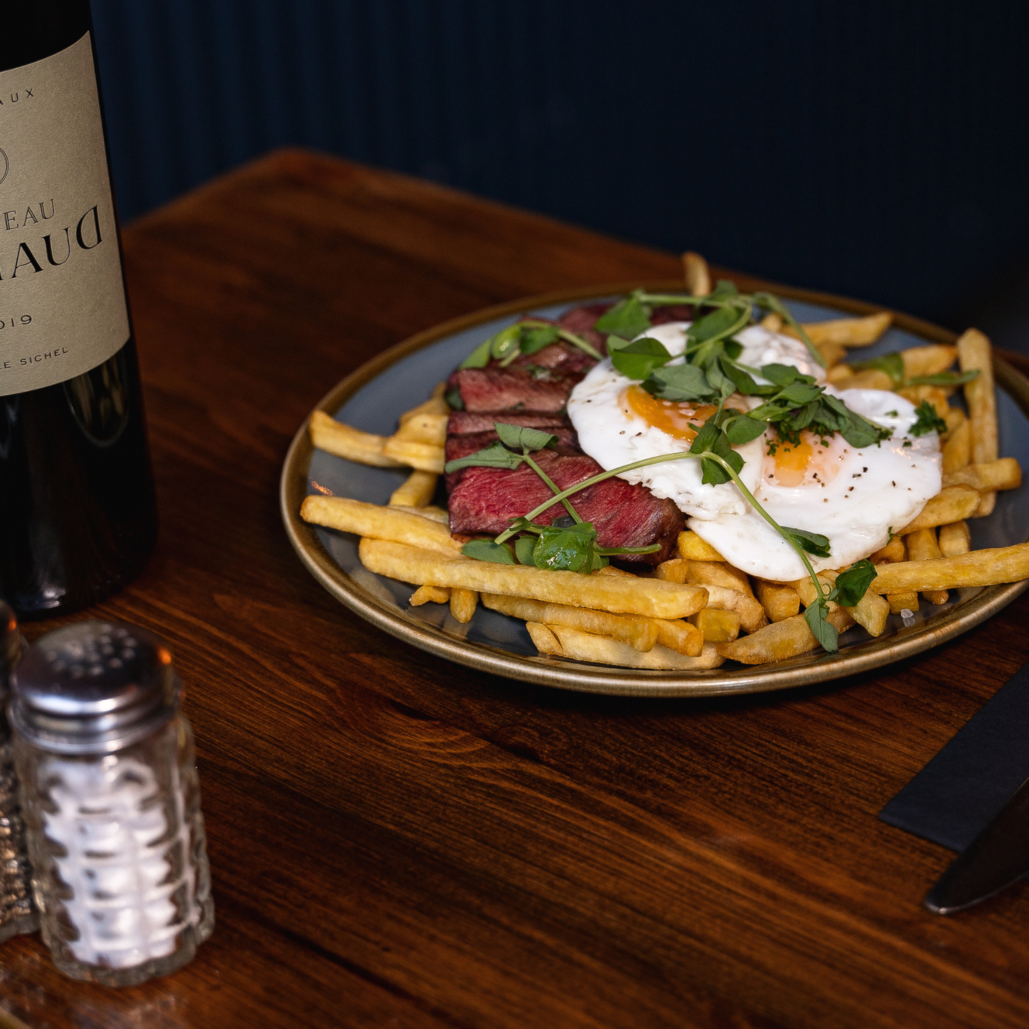 Plate of French fries topped with two fried eggs, beef steak slices, and garnished with green herbs, on a dark dish on a wooden table.