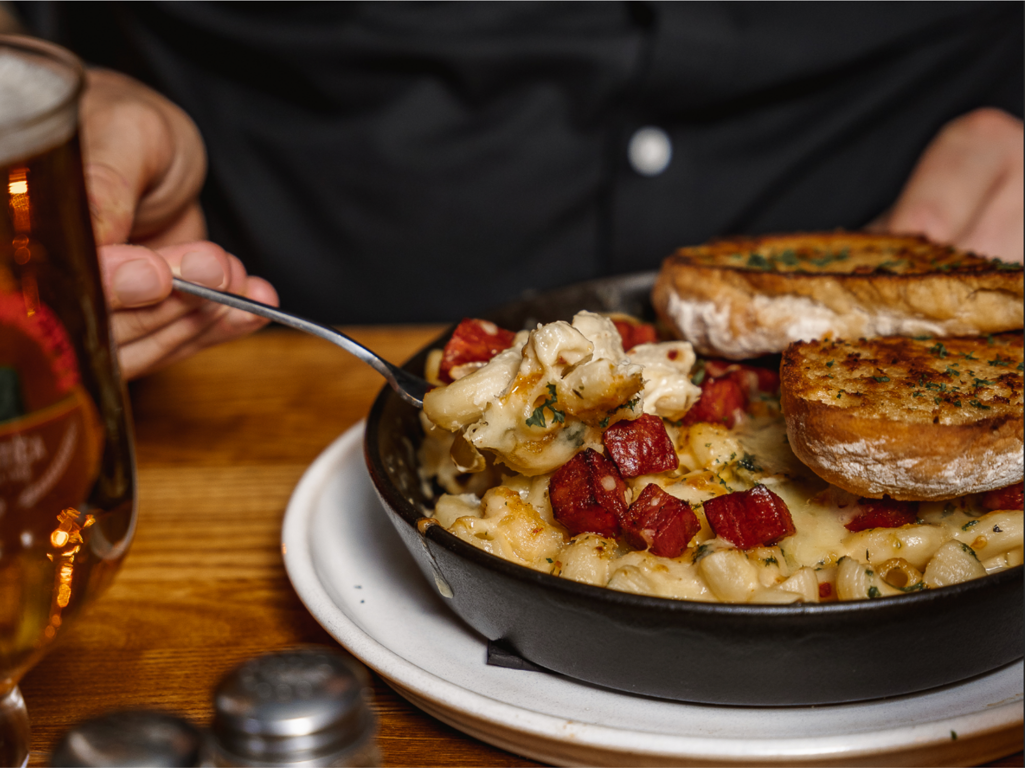A bowl of macaroni and cheese topped with chunks of bacon served with grilled cheese sandwiches on a plate, with a glass of beer in the background.