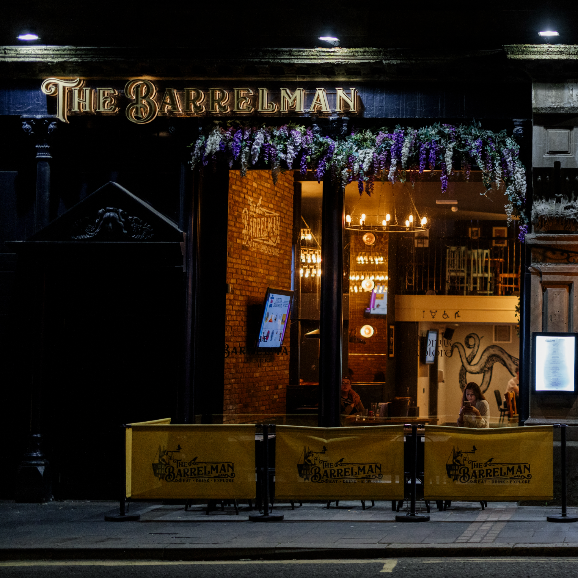 Nighttime exterior view of The Barrelman bar with illuminated sign, decorated with purple and white flowers above the entrance, and patio seating with tables covered with yellow tablecloths, and a lit interior visible through large glass windows.