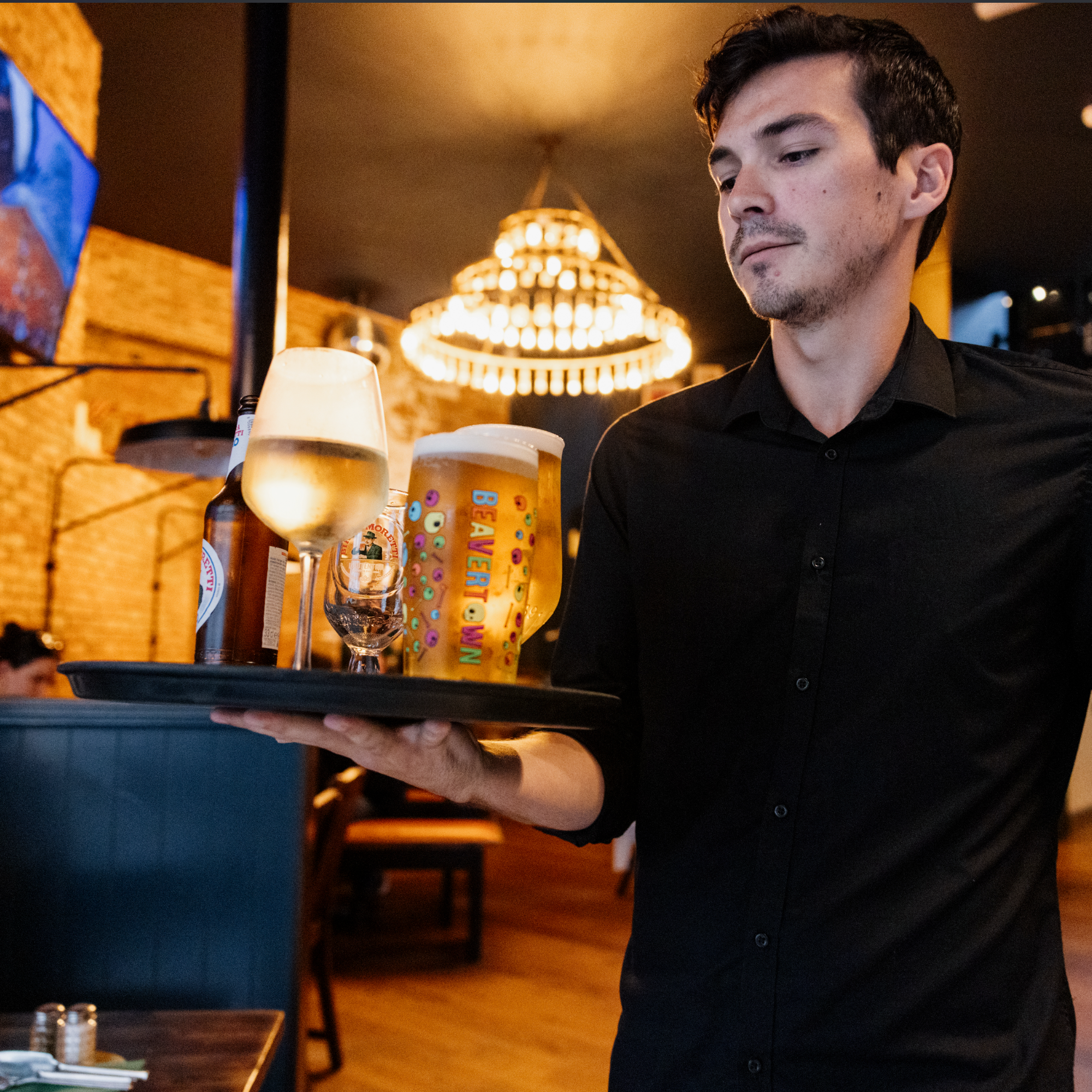 A waiter in a black shirt serving drinks in a restaurant or bar, with a background featuring a illuminated chandelier and warm ambient lighting.
