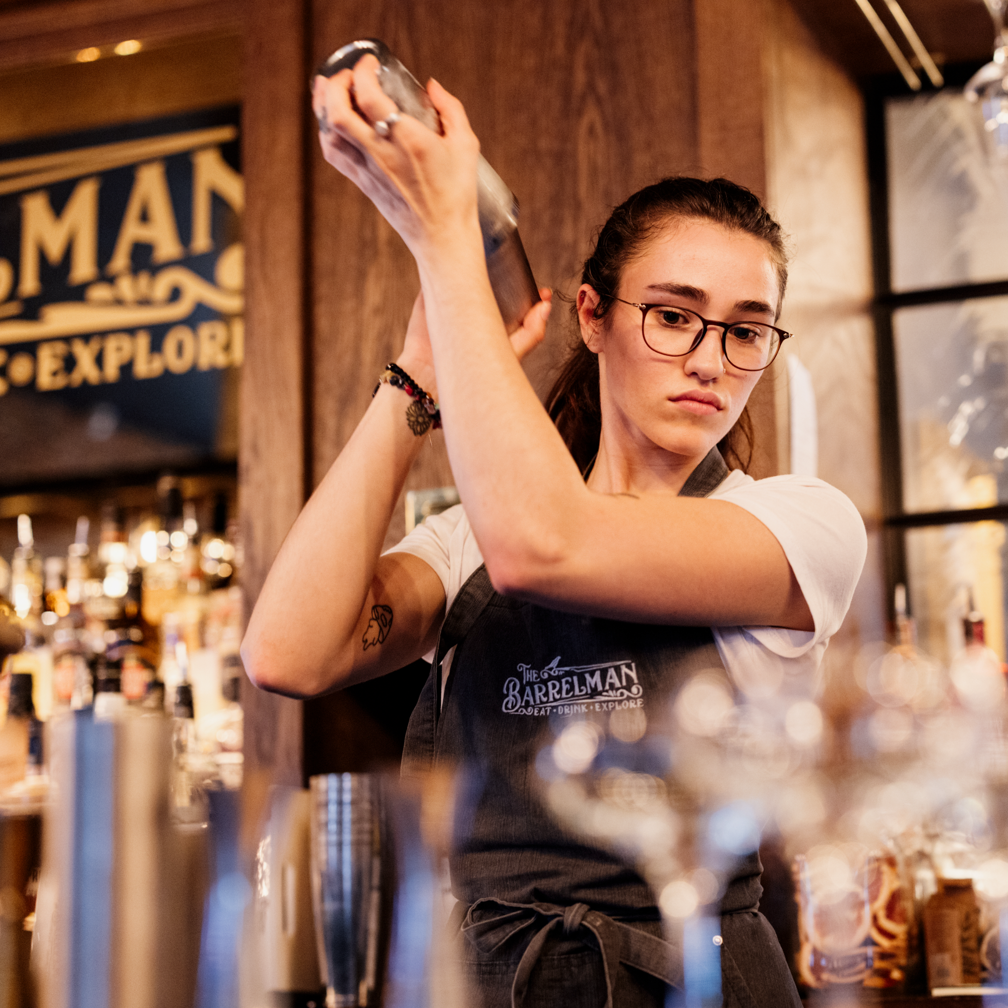 A woman with glasses and a tattoo on her arm is shaking a cocktail shaker behind a bar.