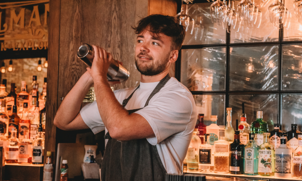 Bartender smiling and holding a cocktail shaker in a bar with various liquor bottles behind him.